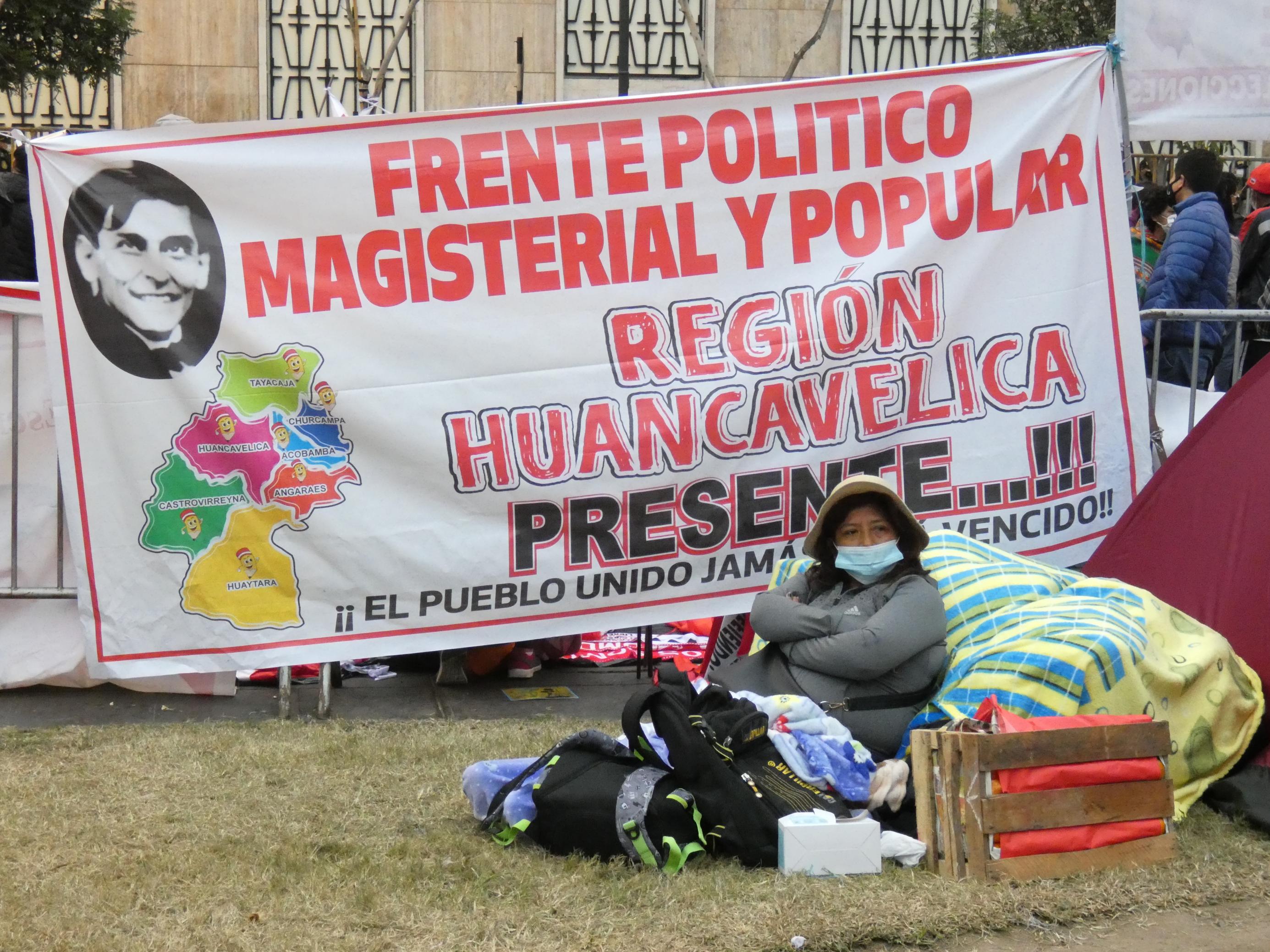 Ein großes Plakat mit der spanischen Aufschrift der Lehrergewerkschaft; davor sitzt eine Person mit viel Gepäck auf dem Rasen.