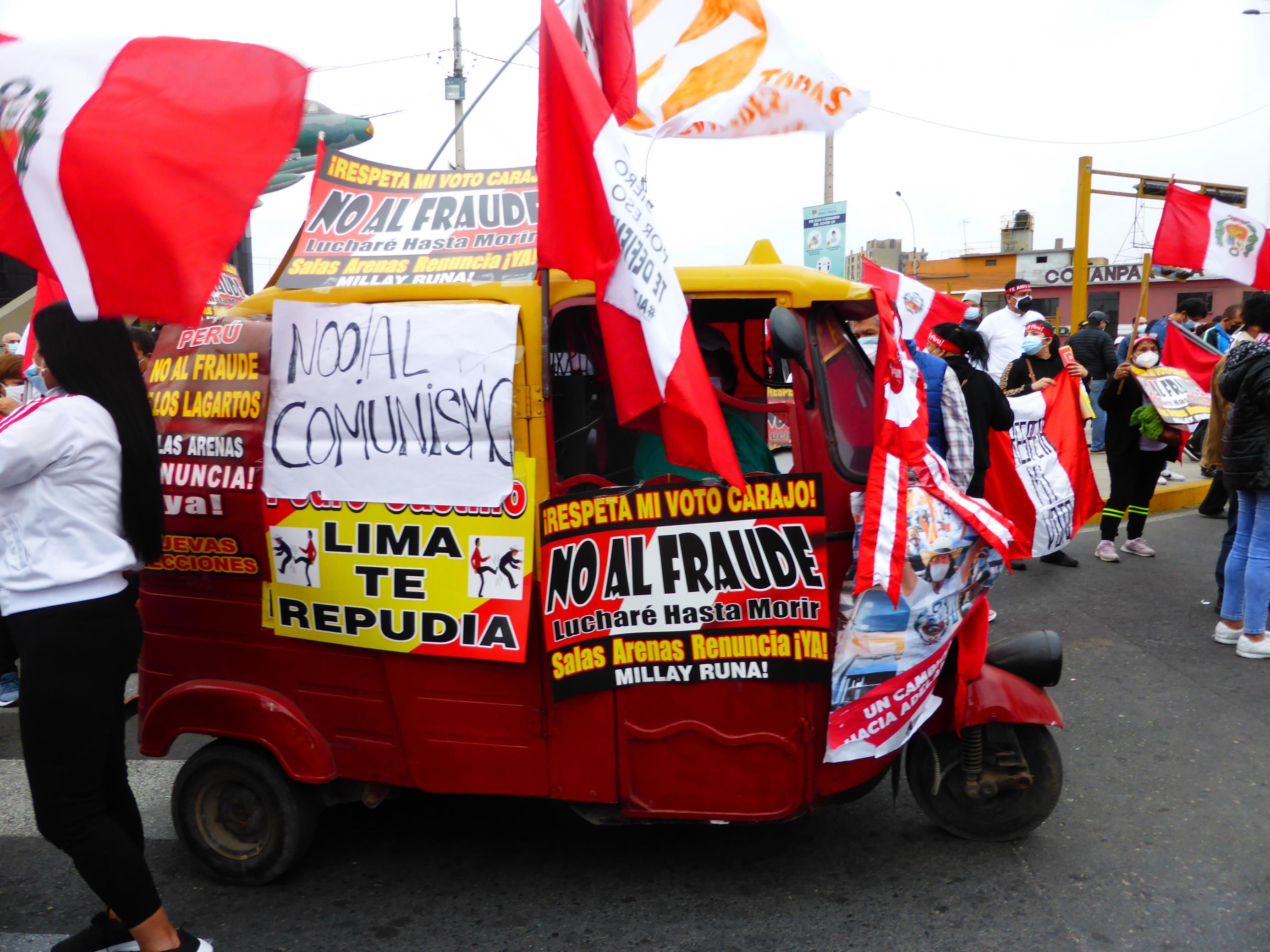 Eine dreirädrige Autorikscha mitten in einer Demo. Personen, die rot-weiße Fahnen schwingen, die Landesfarben Perus.