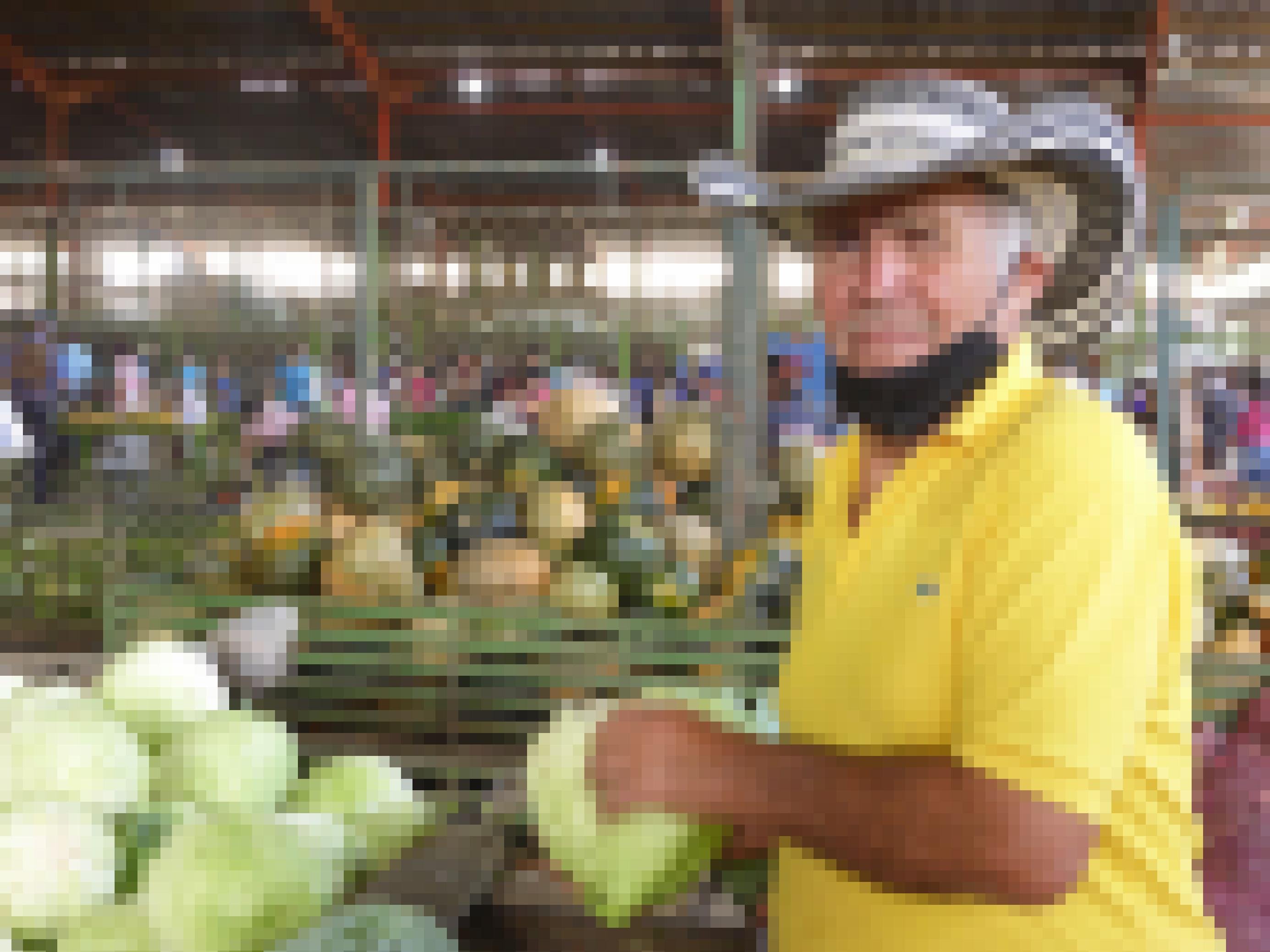 Markthalle mit Gemüseständen. Ein rund 70-jähriger Mann mit Cowboy-Hut und gelbem T-Shirt hält einen Weisskohl in der Hand und blickt in die Kamera.