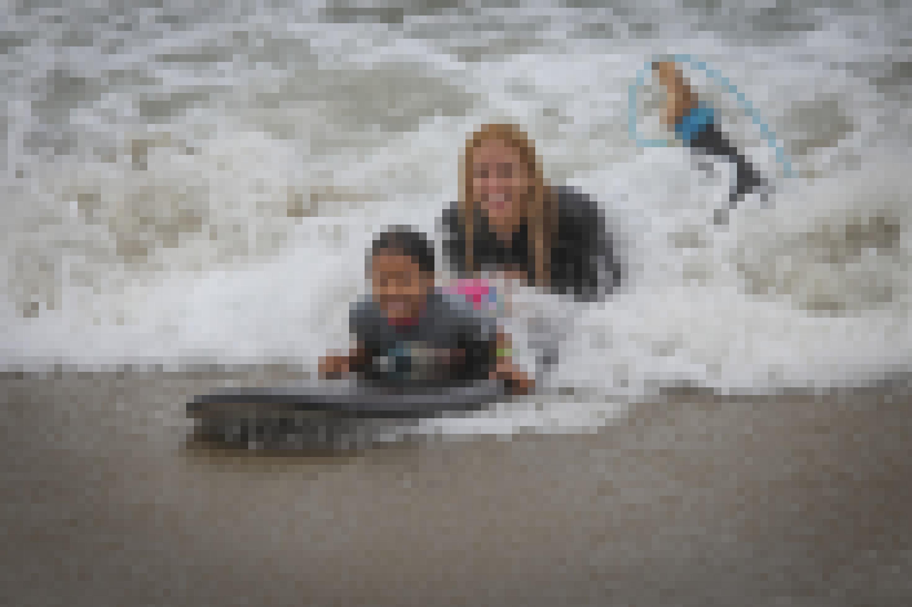 Eine Frau und ein Mädchen lassen sich auf einem Surfbrett von Wellen an den Strand treiben. Beide lachen in die Kamera.