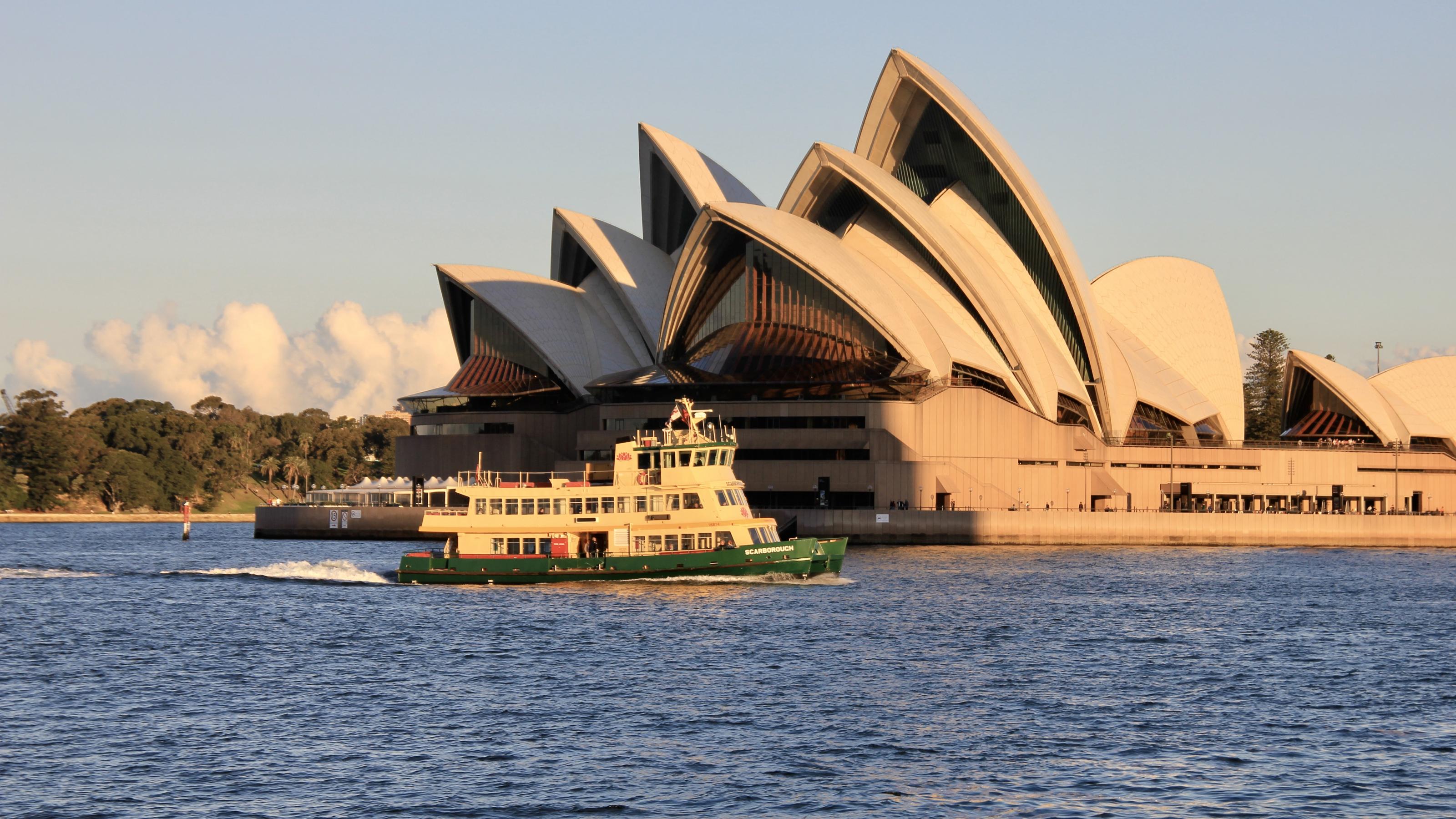 Das muscheförmige weiße Dach des Sydney Opera Houses am Hafen von Sydney. Es ist eines der Wahrzeichen Australiens. Vor dem Gebäude sieht man eine der gelb-grünen Hafenfähren im Wasser.