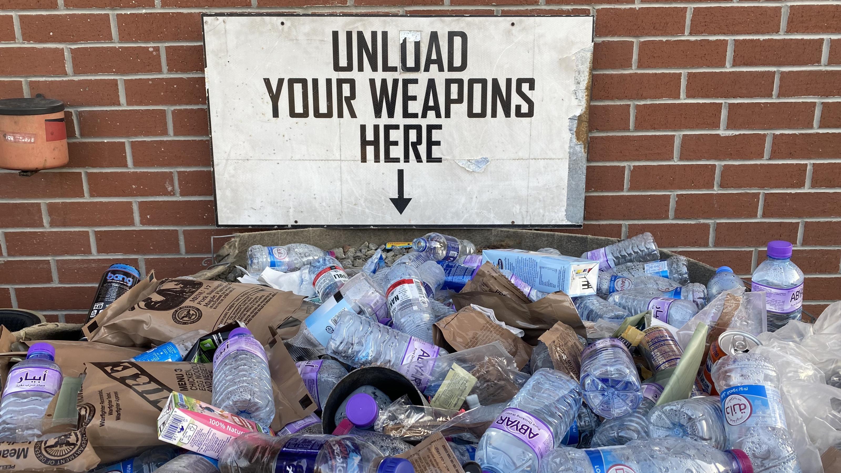 Unter dem Schild mit der Aufschrift „Unload your weapons here“ und der Kiste mit Sand, in die die Patronenhülsen sollen, sind vor allem leere Wasserflaschen zu sehen. Hinweis auf die vielen Zivilisten, die am Flughafen Zuflucht gesucht hatten.