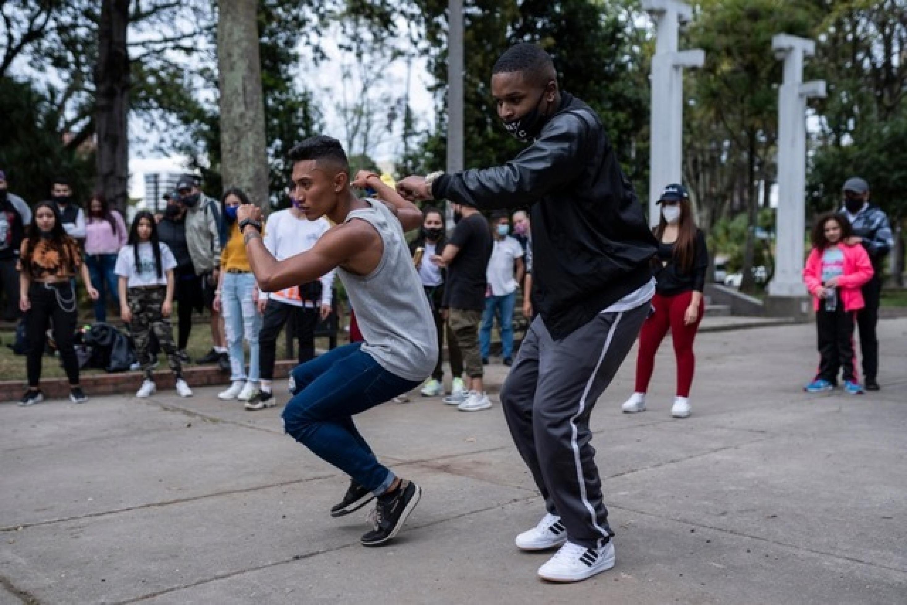 Zwei junge schwarze Männer in Jeans und Traingshose tanzen auf einem öffentlichen Platz. Der eine trägt einen verrutschten Mundschutz, der andere gar keinen. Im Hintergrund sieht man Zuschauer*innen.