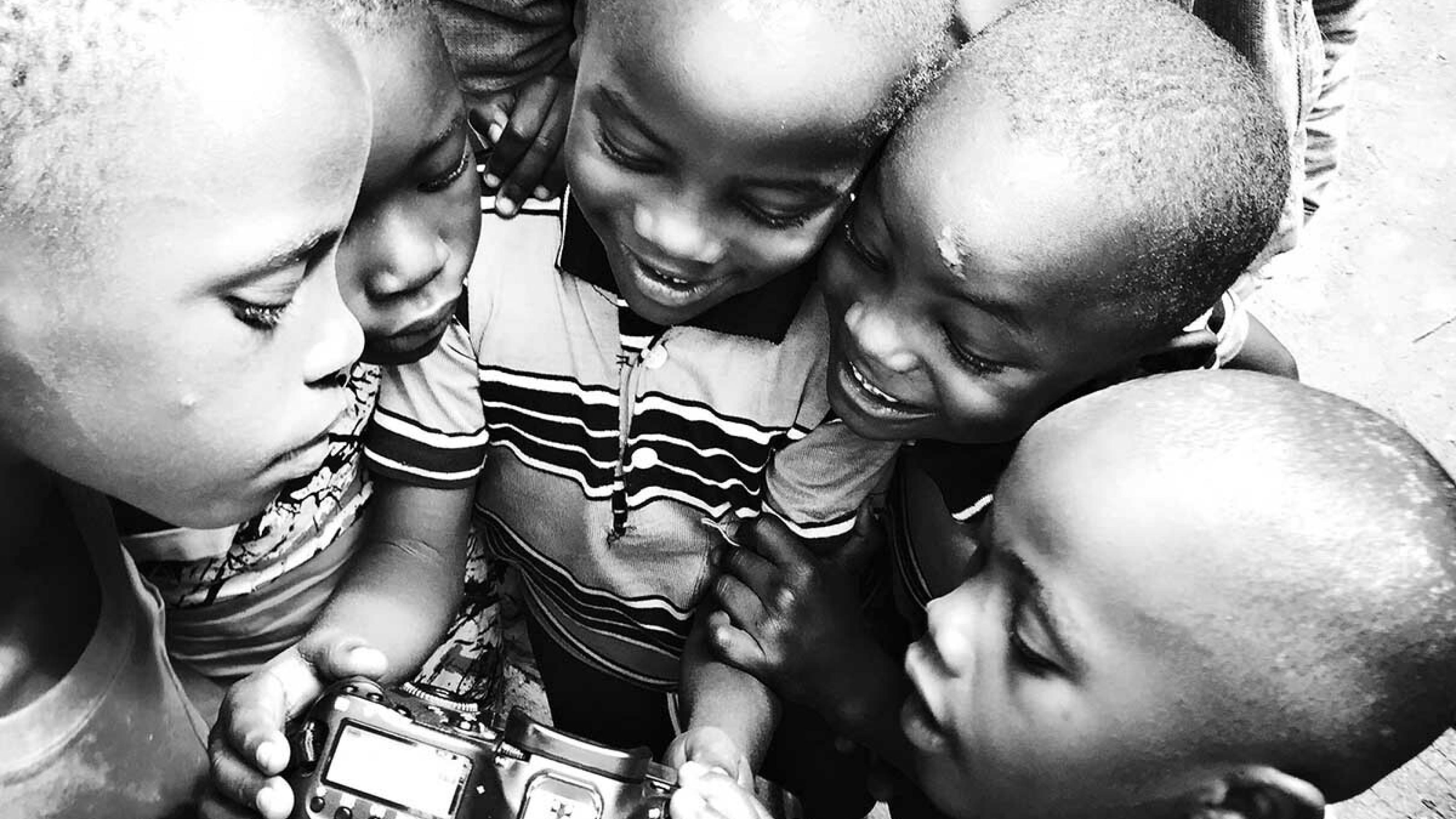5 Kinder stehen kreisförmig um eine DSLR-Kamera herum und betrachten neugierig, was darauf zu sehen ist.