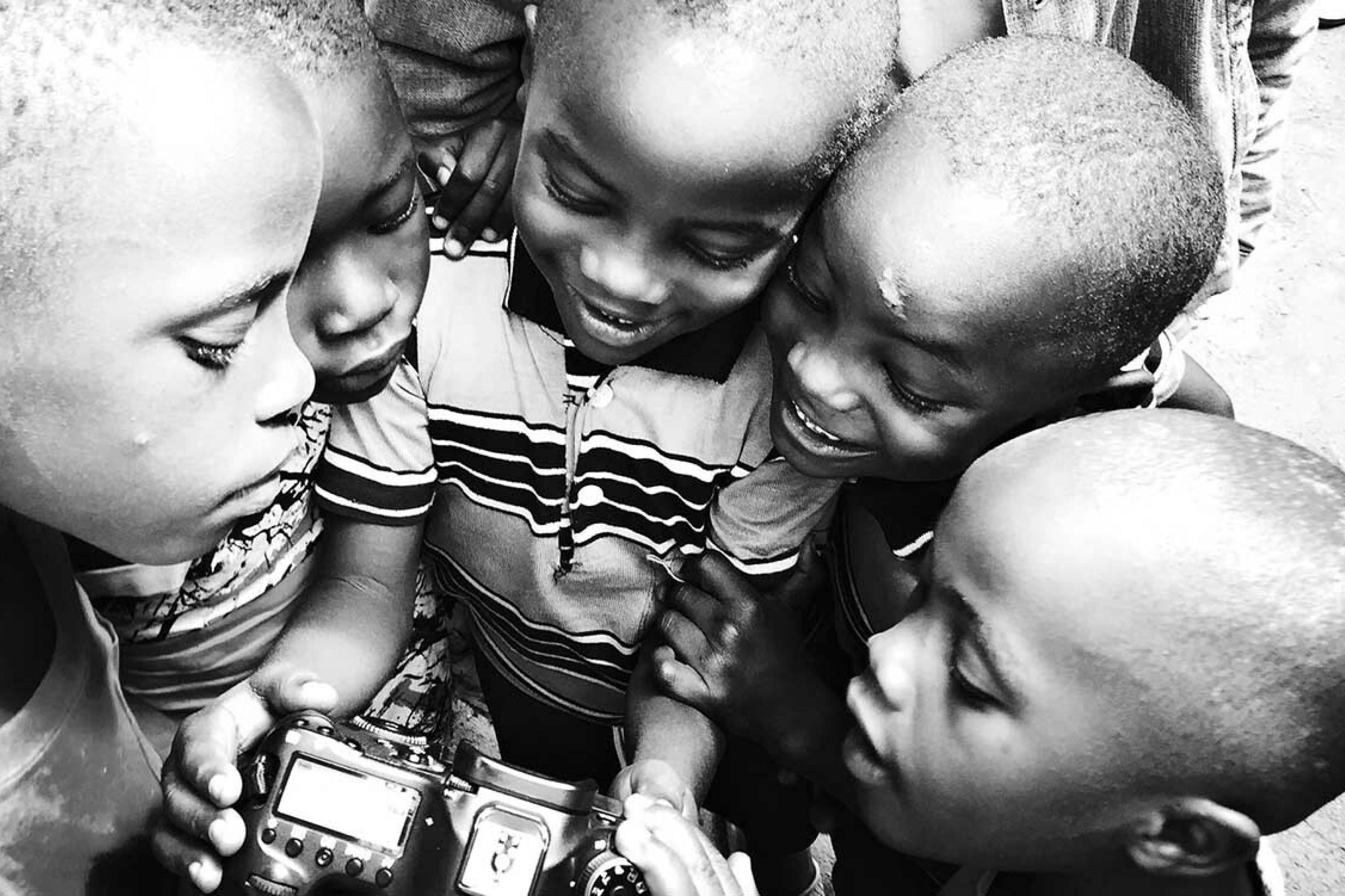 5 Kinder stehen kreisförmig um eine DSLR-Kamera herum und betrachten neugierig, was darauf zu sehen ist.