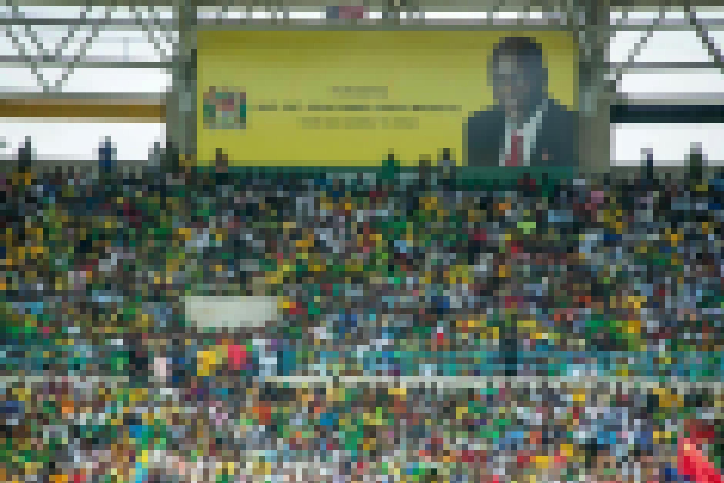 Zu sehen sind die voll besetzten Tribünen in einem Stadion, auf einer riesigen Leinwand ist das Bild des verstorbenen Präsidenten John Magufuli projiziert.