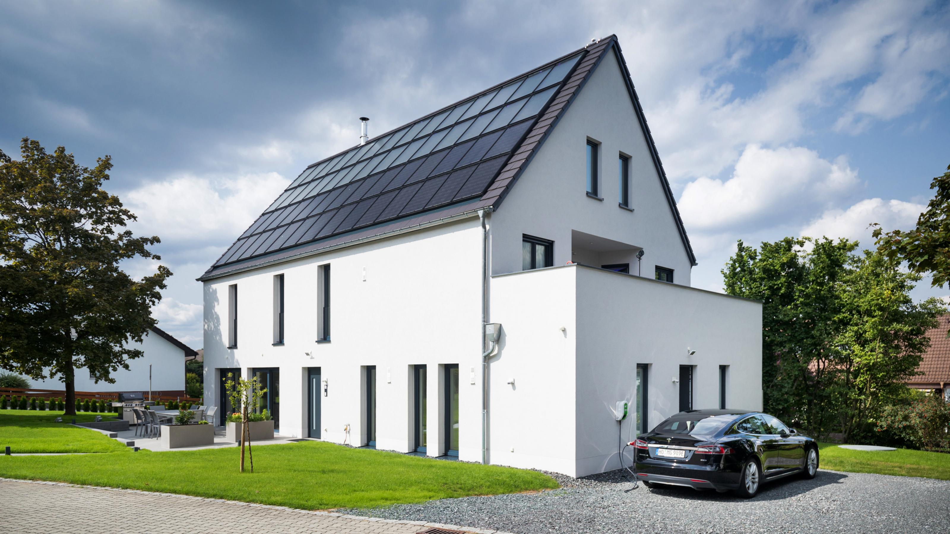 Weiß verputztes, modernes Einfamilienhaus mit Photovoltaik und Solarthermie auf dem Dach, im Vordergrund ein Garten.