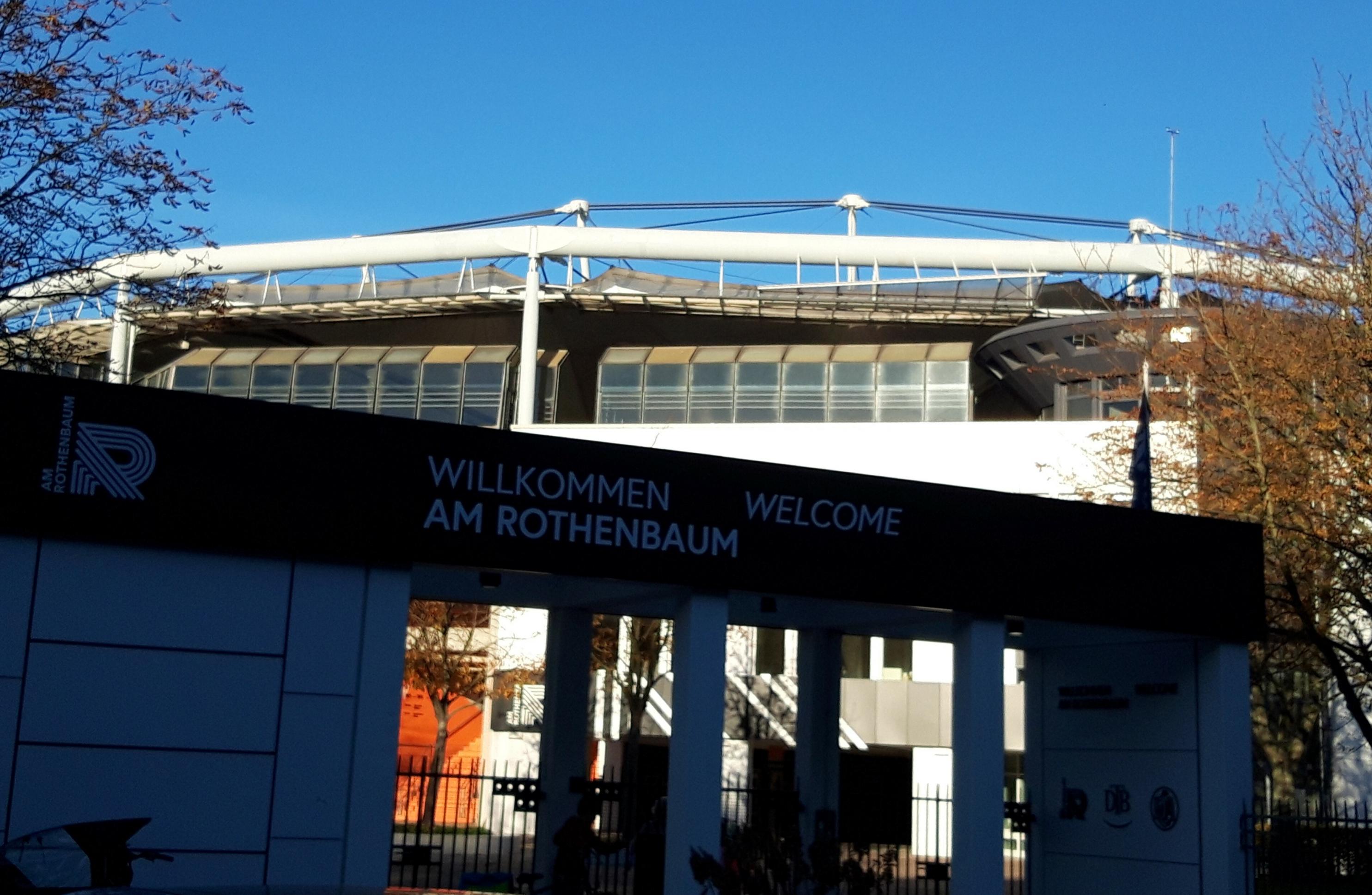 Straßenansicht der Eingangspforte, auf der zu lesen ist „Willkommen am Rothenbaum“ – letzteres ebenso zusammengeschrieben wie die gleich weiter westlich des im Hintergrund aufragenden Stadions gelegene Straße namens Rothenbaumchaussee.