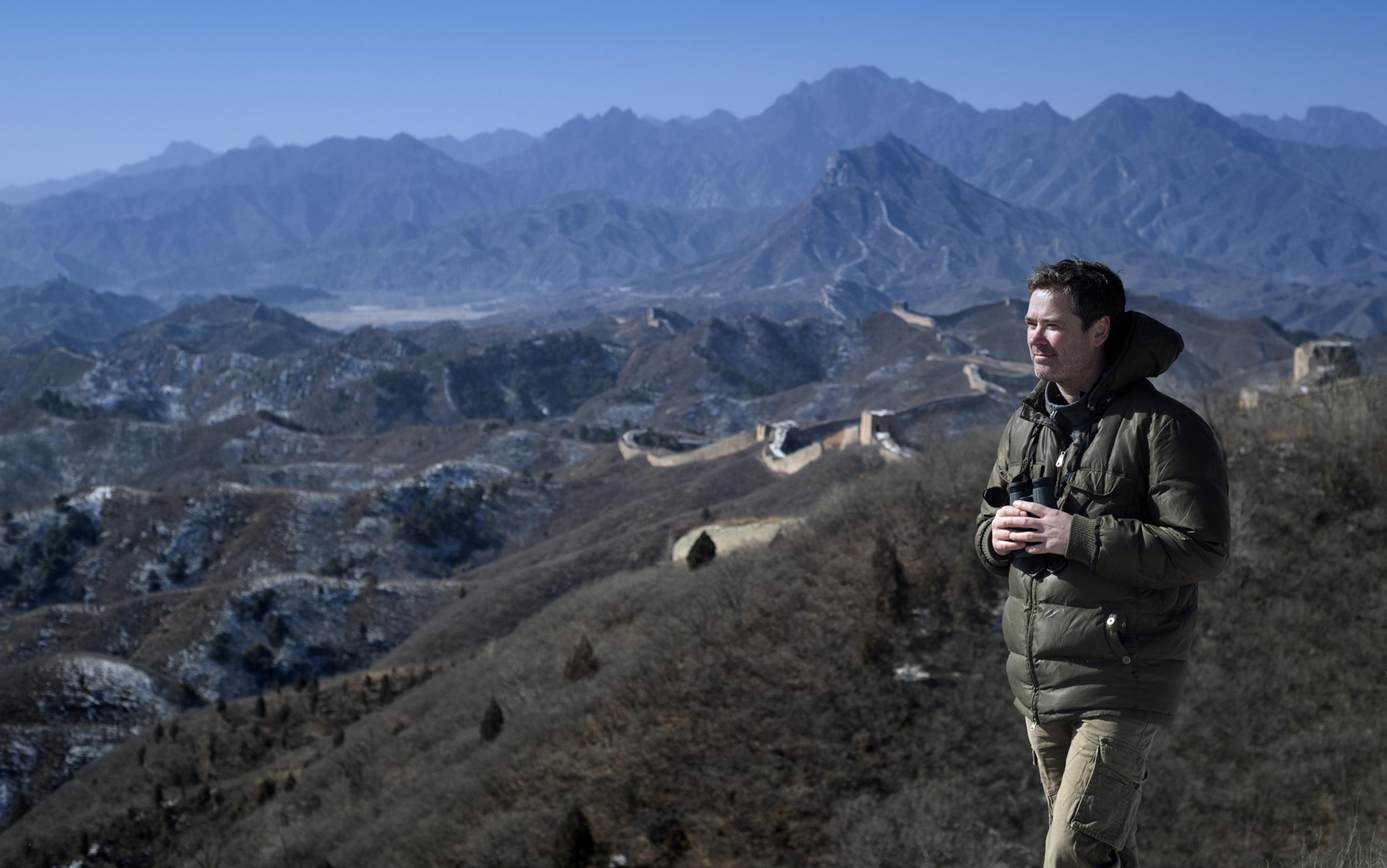 Auf dem Bild ist Terry Townsend, unser Interviewpartner, zu sehen, wie er vor der Großen Mauer in China steht und ein Fernglas in der Hand hält.