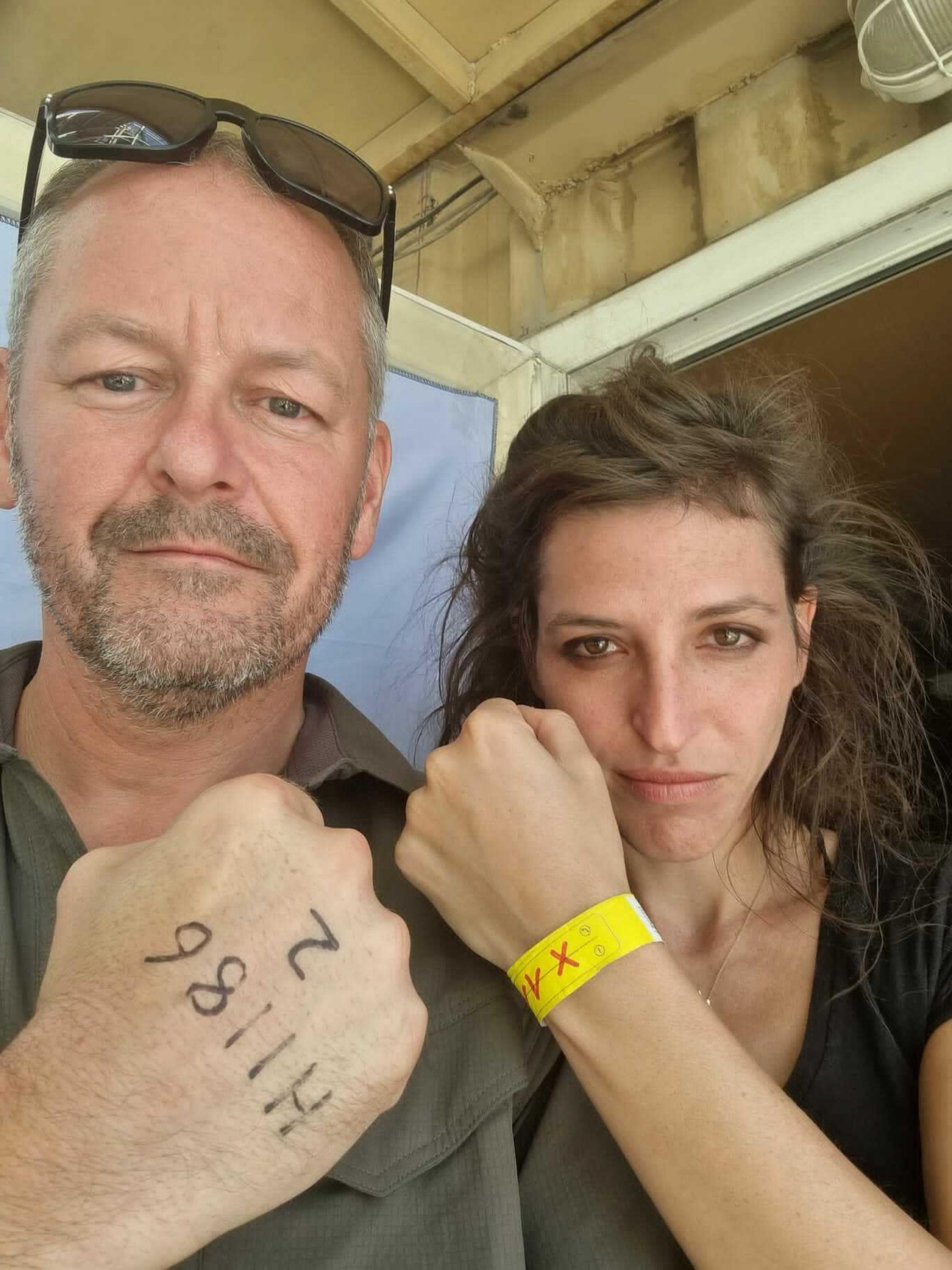 Rob Grey und Theresa Breuer halten beide ihre Handgelenke hoch. Grey hat das britische Evakuierungskennzeichen auf dem Handrücken stehen, Breuer das deutsche auf einem gelben Armband.