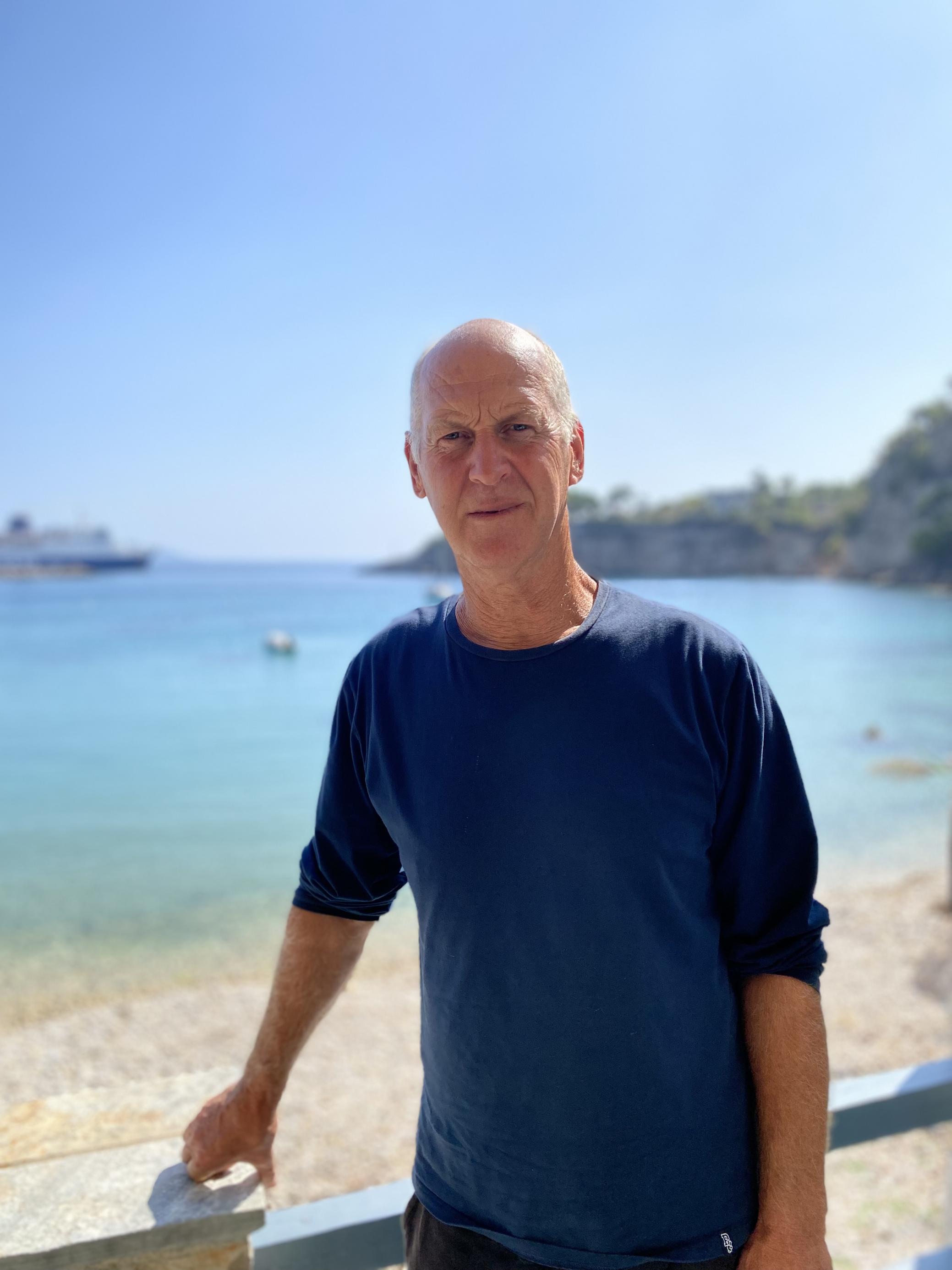 Gut gebräunter Mann mit Glatze und blauem Sweatshirt steht vor dem türkisen Meer und schaut nachdenklich in die Kamera