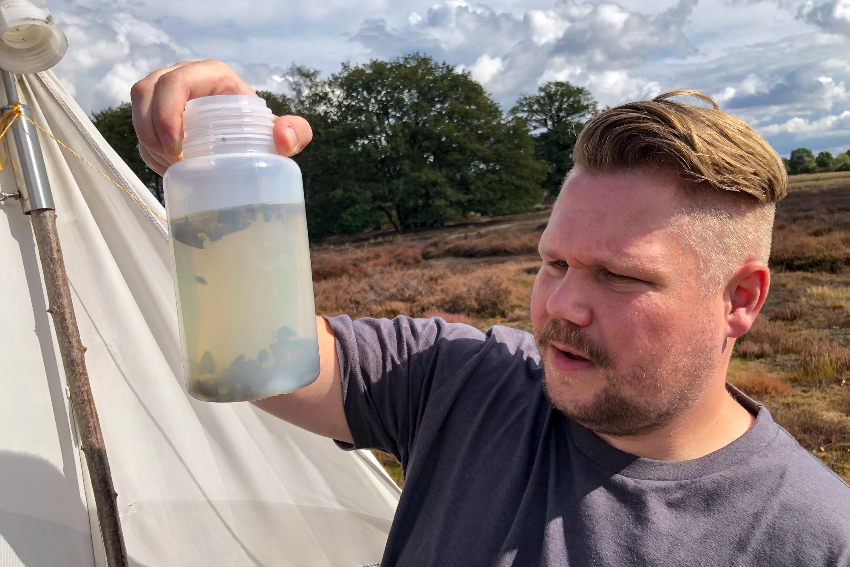 Ein Mann hält eine Plastikflasche, in der Insekten in einer Flüssigkeit schwimmen, hoch und schaut sie an.