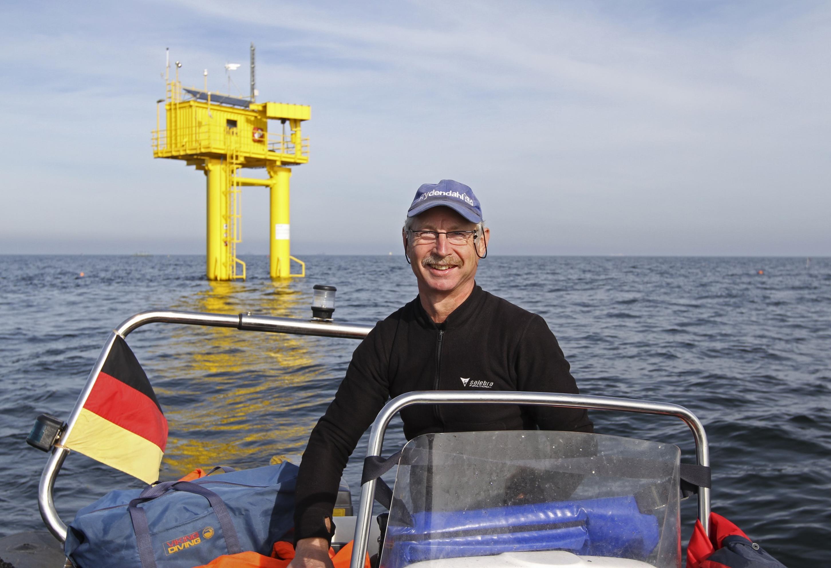 Älterer Mann auf einem Boot auf dem Meer, im Hintergrund eine gelbe Plattform, die aus dem Wasser ragt