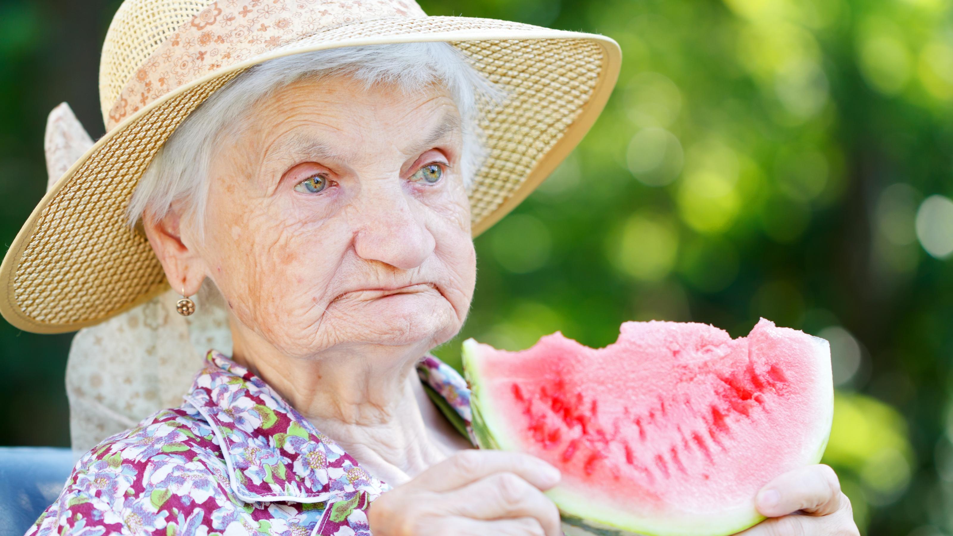 Eine alte Dame mit Hut hält eine Scheibe Wassermelone, von der sie bereits abgebissen hat. Obst ist wichtig, um den Flüssigkeitsbedarf zu decken.