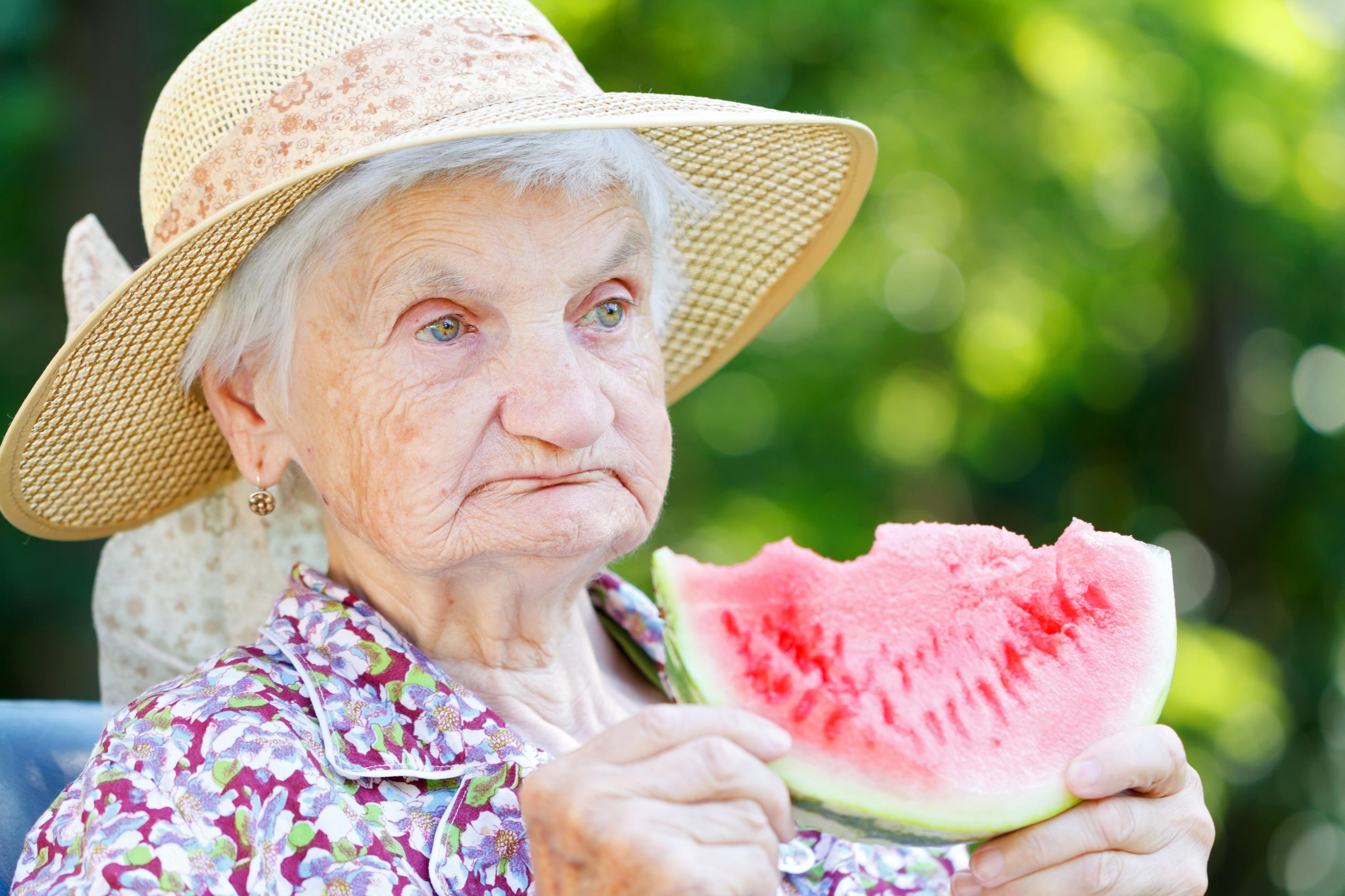 Eine alte Dame mit Hut hält eine Scheibe Wassermelone, von der sie bereits abgebissen hat. Obst ist wichtig, um den Flüssigkeitsbedarf zu decken.