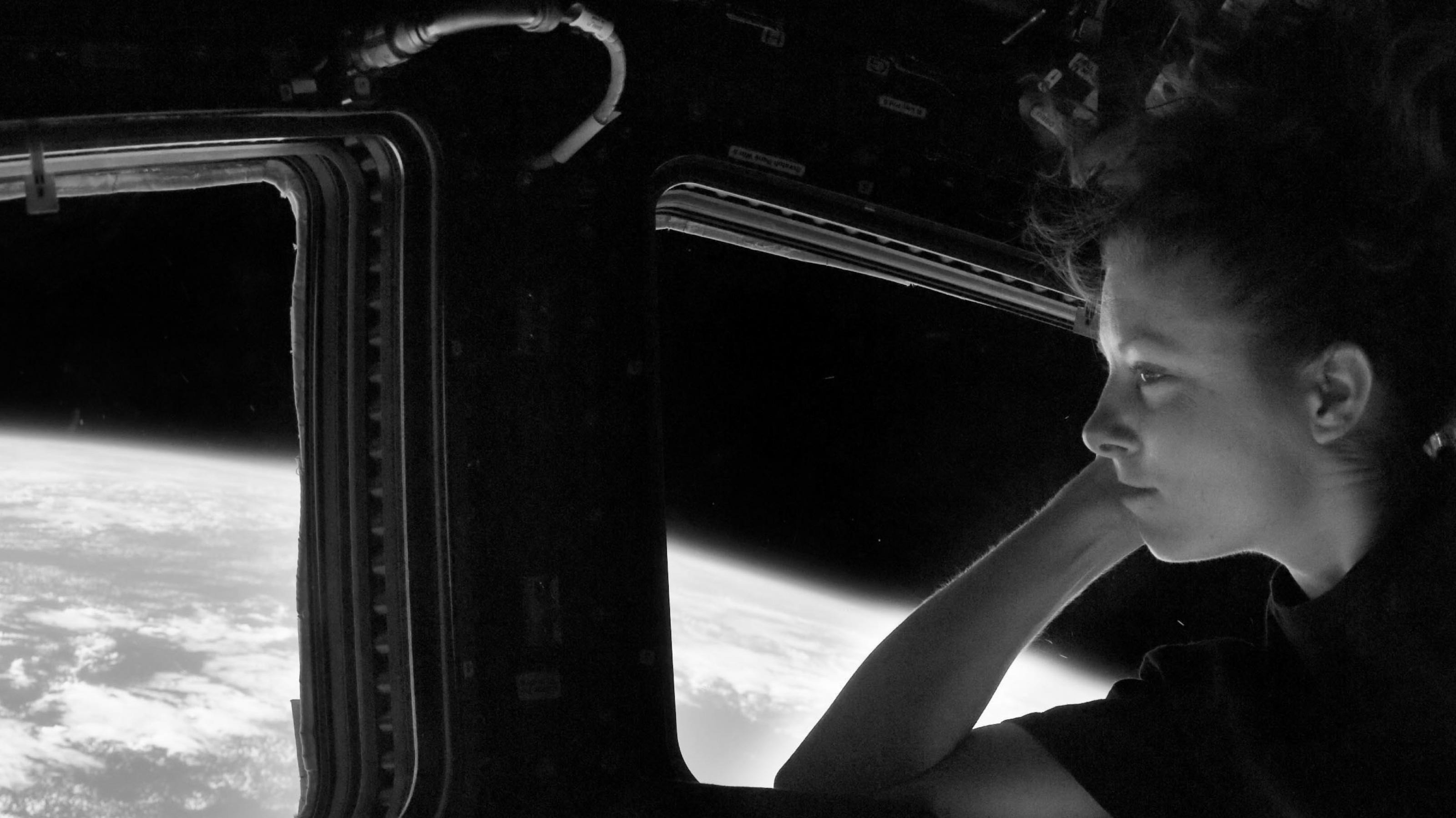 Eine NASA-Astronautin in schwarzem T-Shirt schaut aus einem Fenster der Raumstation und tut so, als würde sie ihren Kopf auf den Arm aufstützen (es sieht sehr entspannt aus).