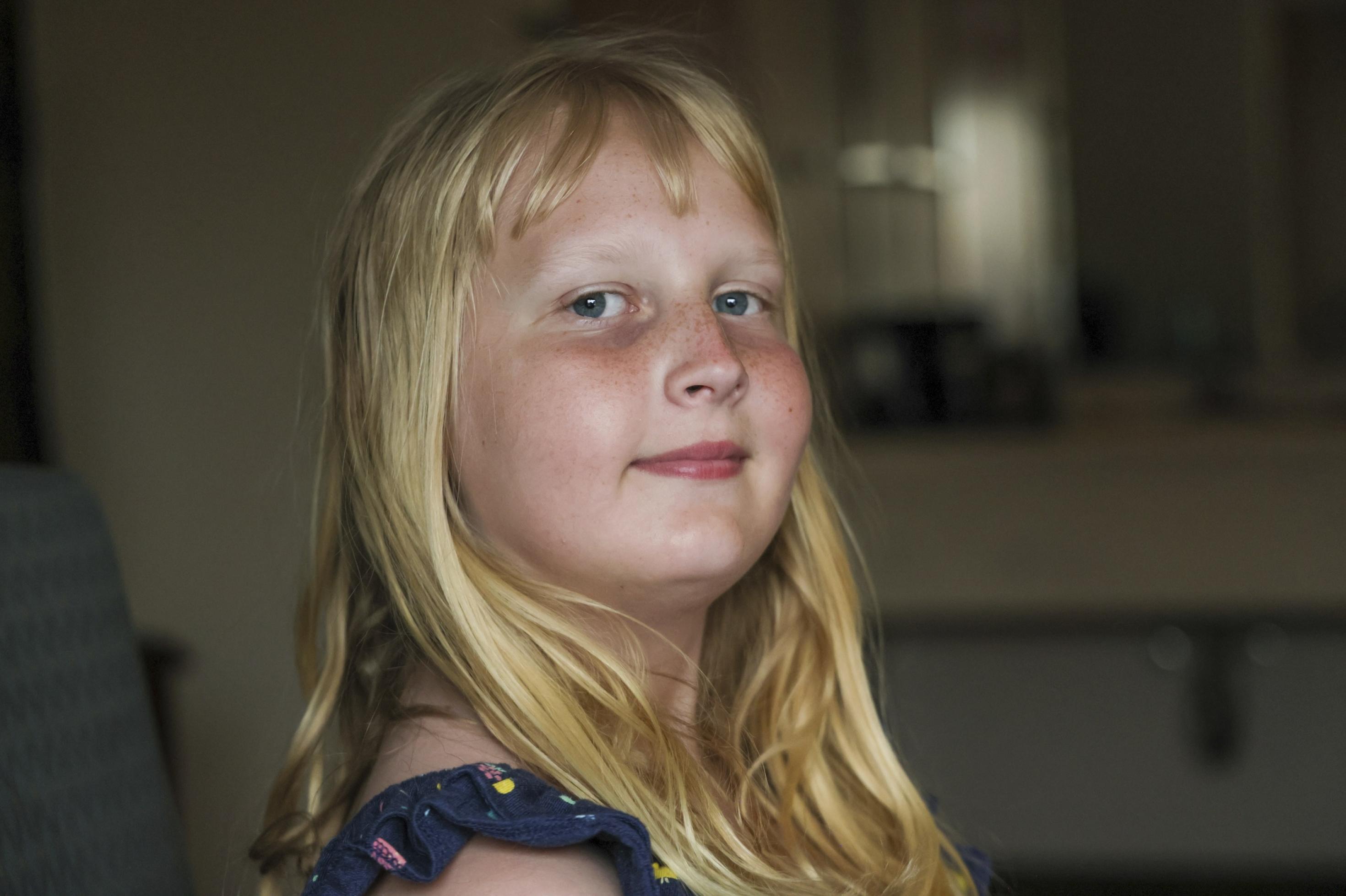 Ein Mädchen mit blauen Augen und langen blonden Haaren blickt selbstbewusst in die Kamera.