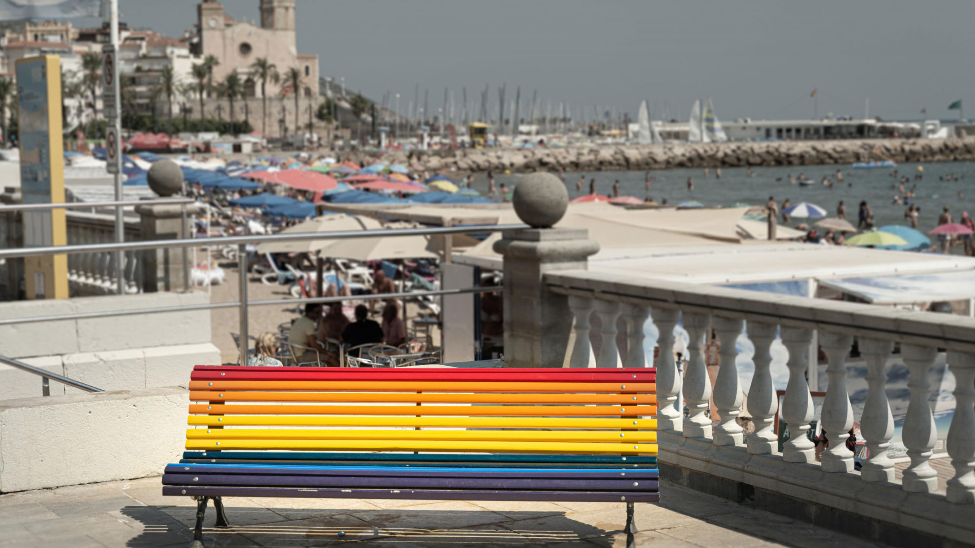 Eine in den Regenbogenfarben bemalte Sitzbank im spanischen Küstenstädtchen Sitges. Im Hintergrund ist eine Kirche, die Strandpromenade und das Meer zu sehen.