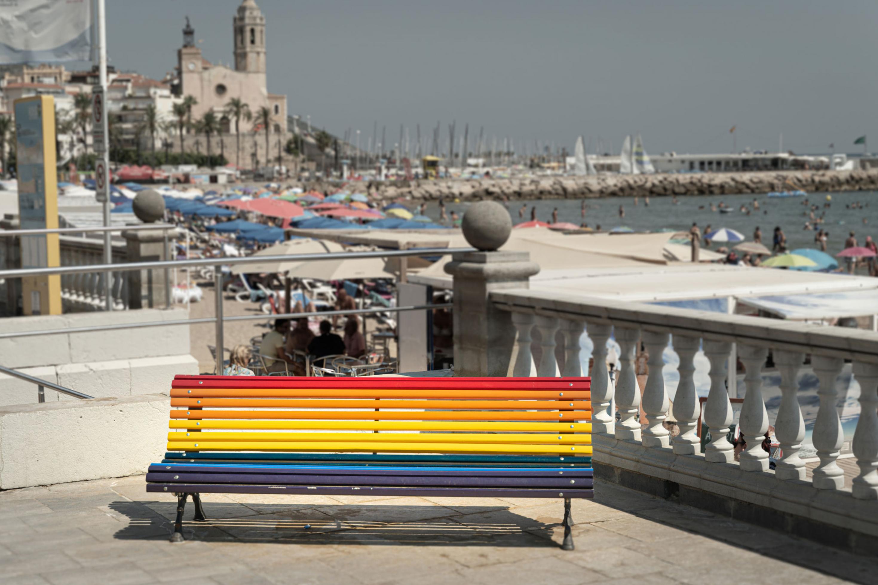 Eine in den Regenbogenfarben bemalte Sitzbank im spanischen Küstenstädtchen Sitges. Im Hintergrund ist eine Kirche, die Strandpromenade und das Meer zu sehen.