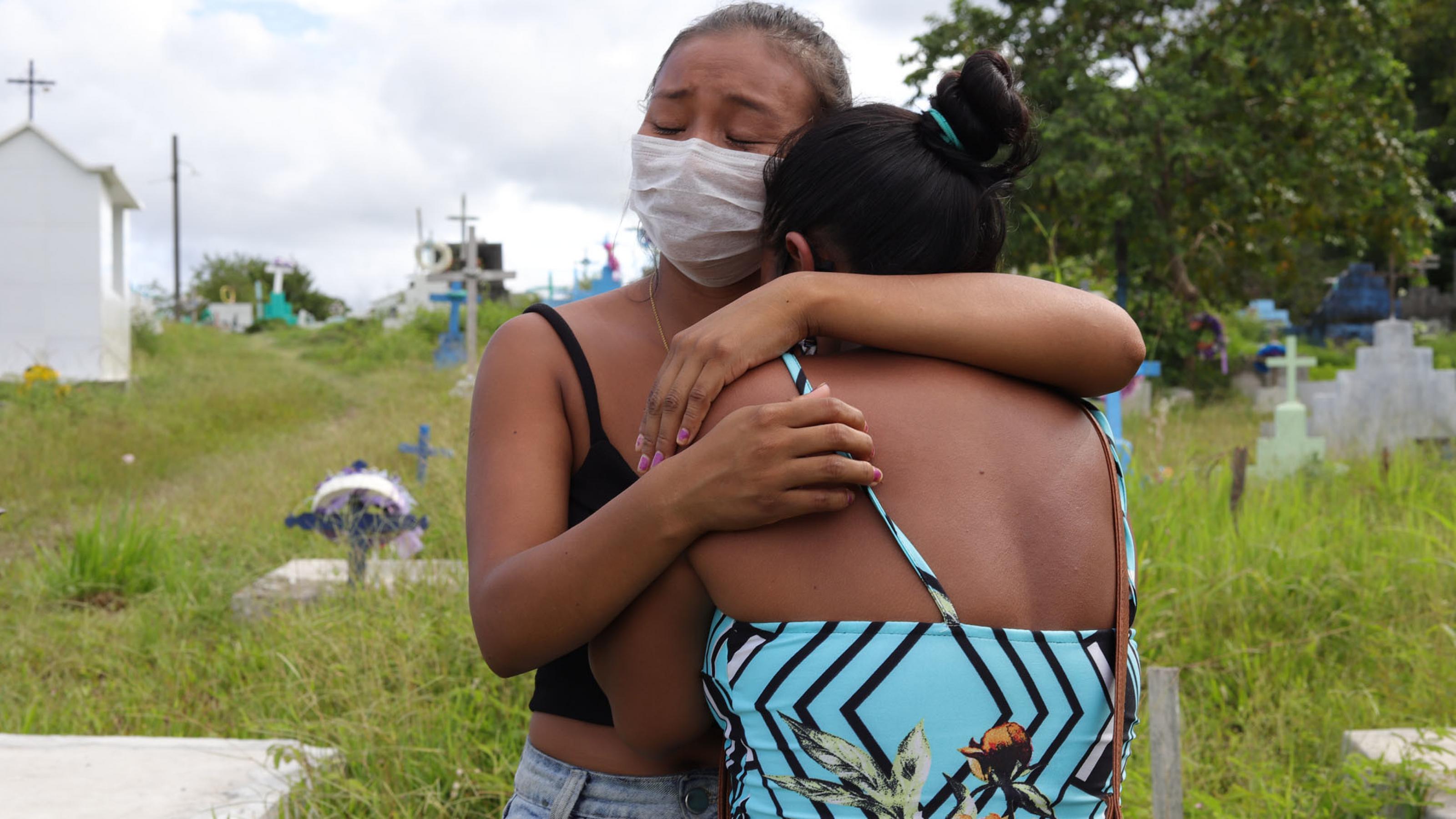 Zwei Frauen trauernde umarmen und trösten sich. Auf dem Friedhof Parque da Saudade, in São Gabriel da Cachoeira im Bundesstaat Amazonas sieht man bunte Kreuze im Hintergrund von hohen Gräsern.