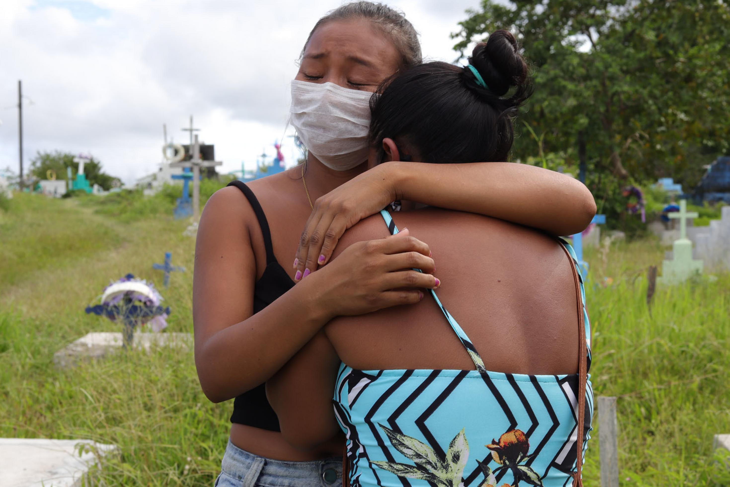 Zwei Frauen trauernde umarmen und trösten sich. Auf dem Friedhof Parque da Saudade, in São Gabriel da Cachoeira im Bundesstaat Amazonas sieht man bunte Kreuze im Hintergrund von hohen Gräsern.