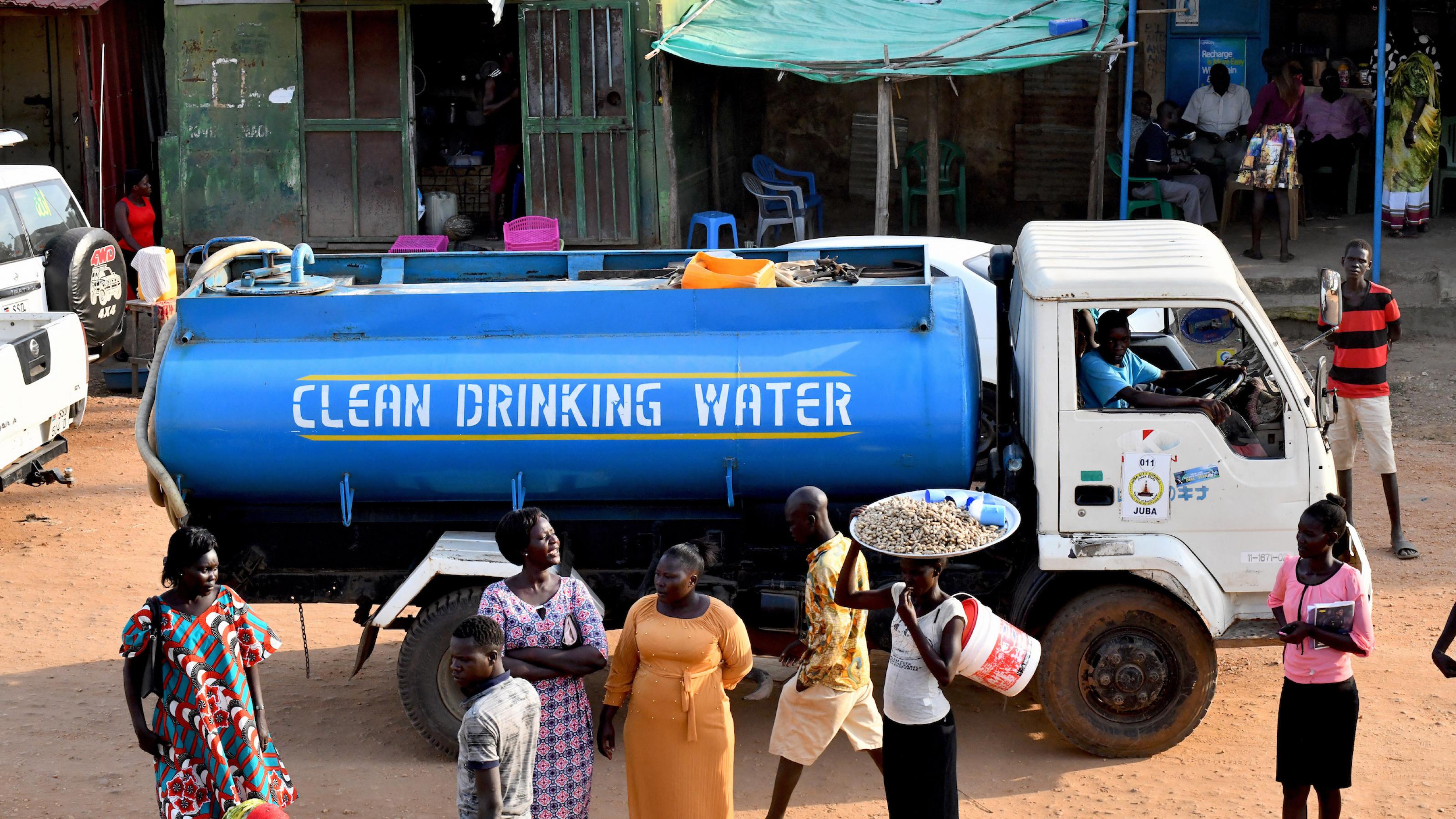 Ein Tanklastwagen fährt auf einer sandigen Straße durch die südsudanesiche Stadt Dschuba. Der Tank trägt die Aufschrift „Clean Drinking Water“, sauberes Trinkwasser. Vor dem LKW stehen Passantinnen.