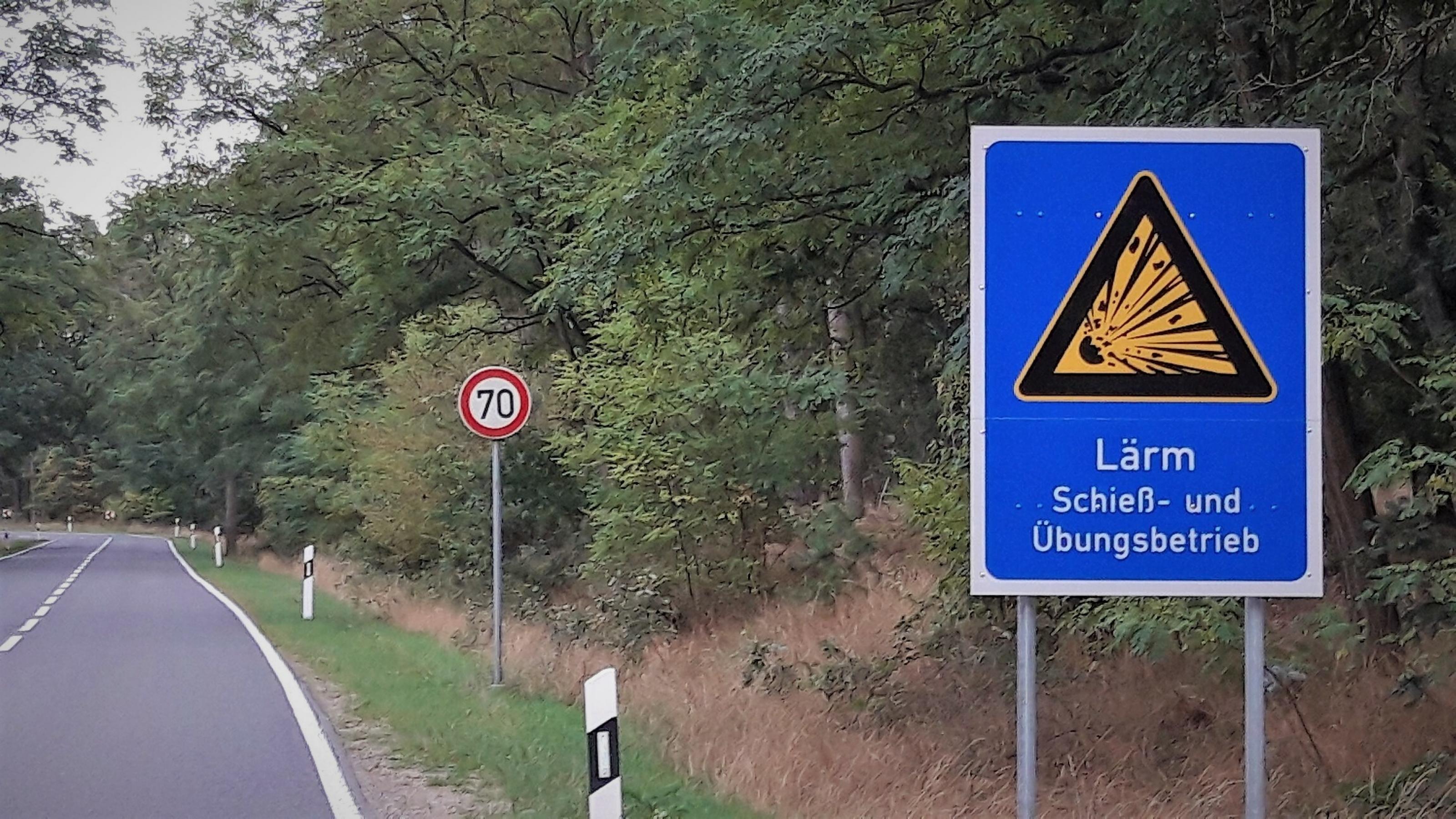 Vor einer waldumsäumten Straße steht das blaue Warnschild mit der Aufschrift „LÄRM – Schieß- und Übungsbetrieb“.