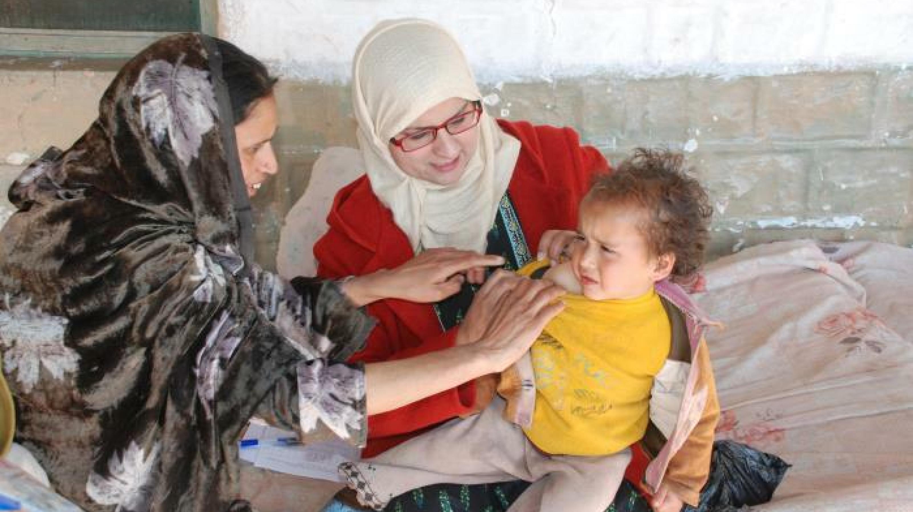 Eine Mutter hält ein Kleinkind in den Armen, eine Frau, eine Ärztin untersucht das Kind nach einer Impfung gegen Tuberkulose.