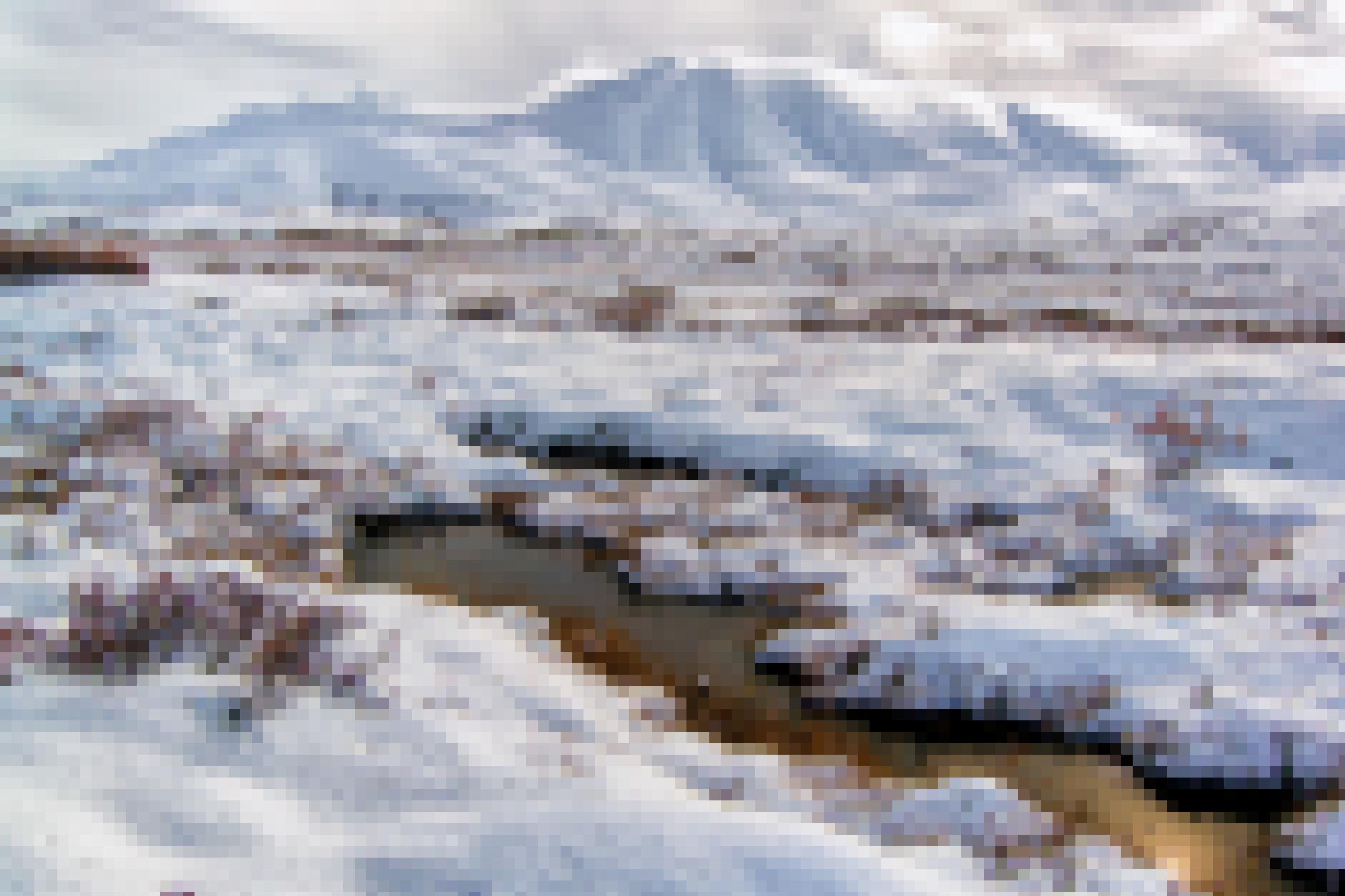 Das Foto zeigt eine schneebedeckte Landschaft, aus der niedrige Sträucher sowie Gräser herausragen. Vorne sind Tümpel mit offenen Wasserflächen zu erkennen, dahinter folgt eine Ebene und am Horizont erheben sich komplett von Schnee bedeckte Berge. Die Sonne scheint von rechts oben auf die Landschaft und glitzert im Wasser der Tümpel.