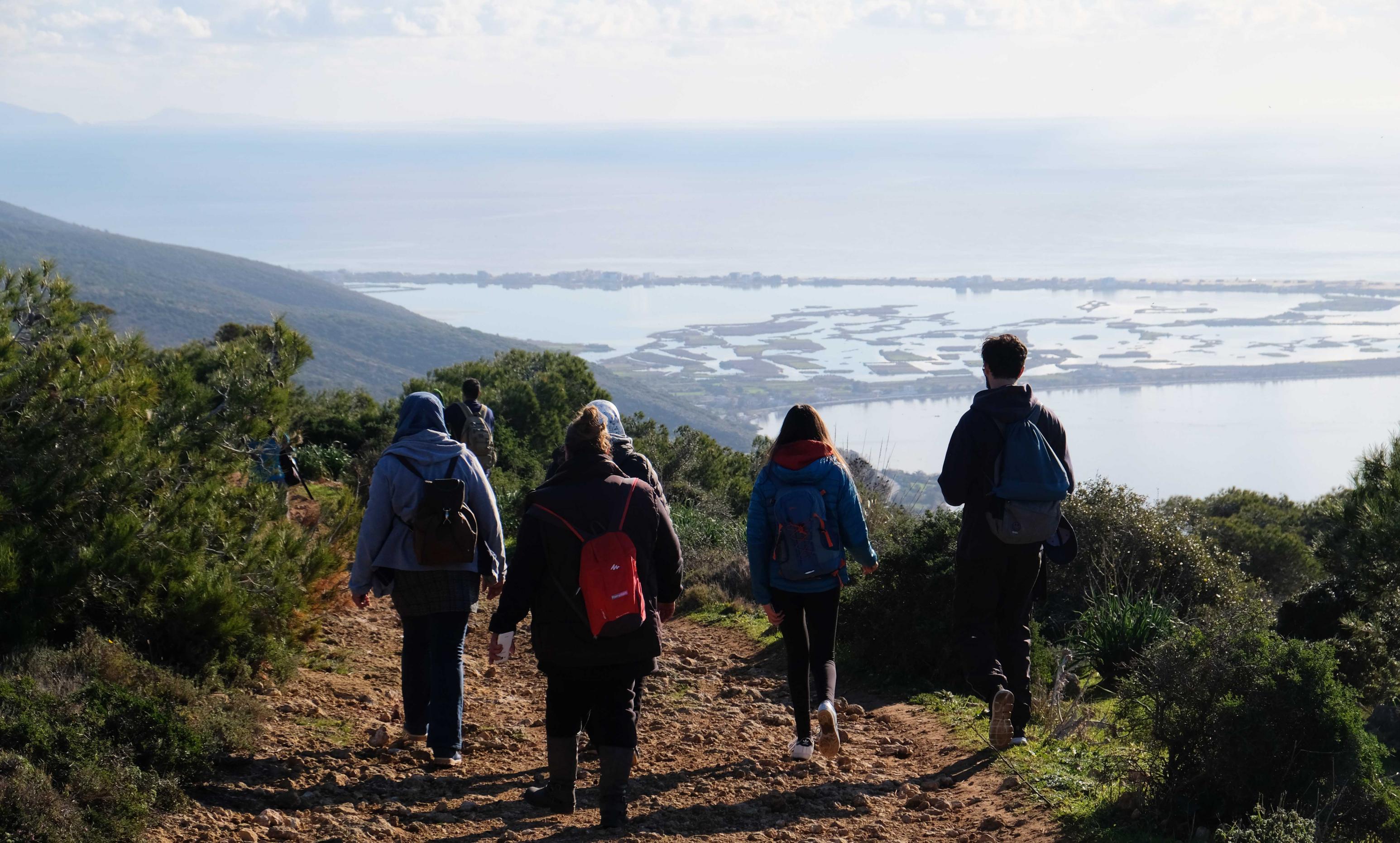 Eine Gruppe junger Menschen in bunter Wanderbekleidung läuft einen Hügel hinunter. Sie sind von hinten zu sehen, vor ihnen das Meer