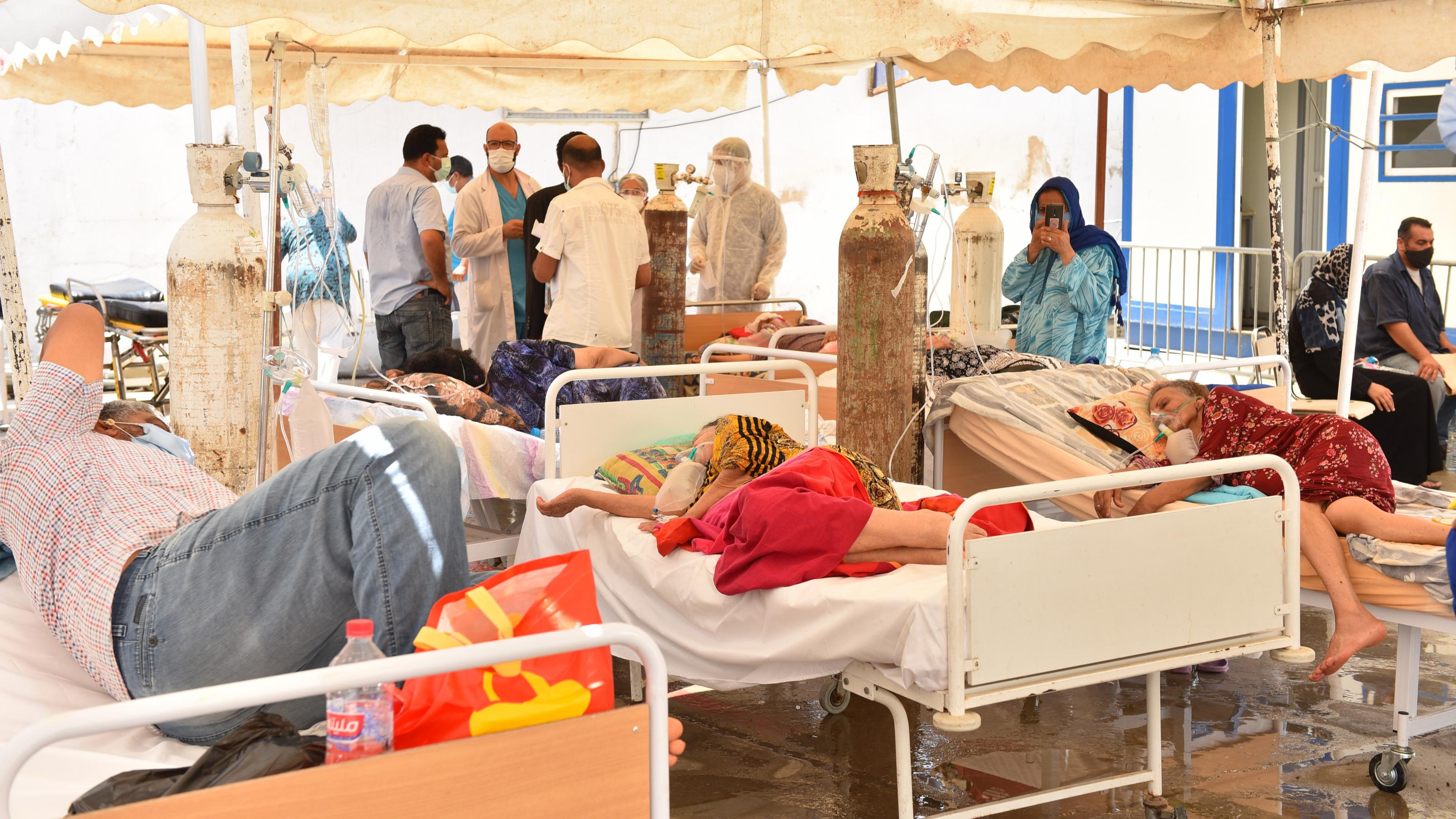 Erkrankte liegen an Sauerstoffflaschen unter einem improvisierten Zelt. Im Hintergrund diskutiert eine Gruppe von Ärztïnnen und Pflegekräften.