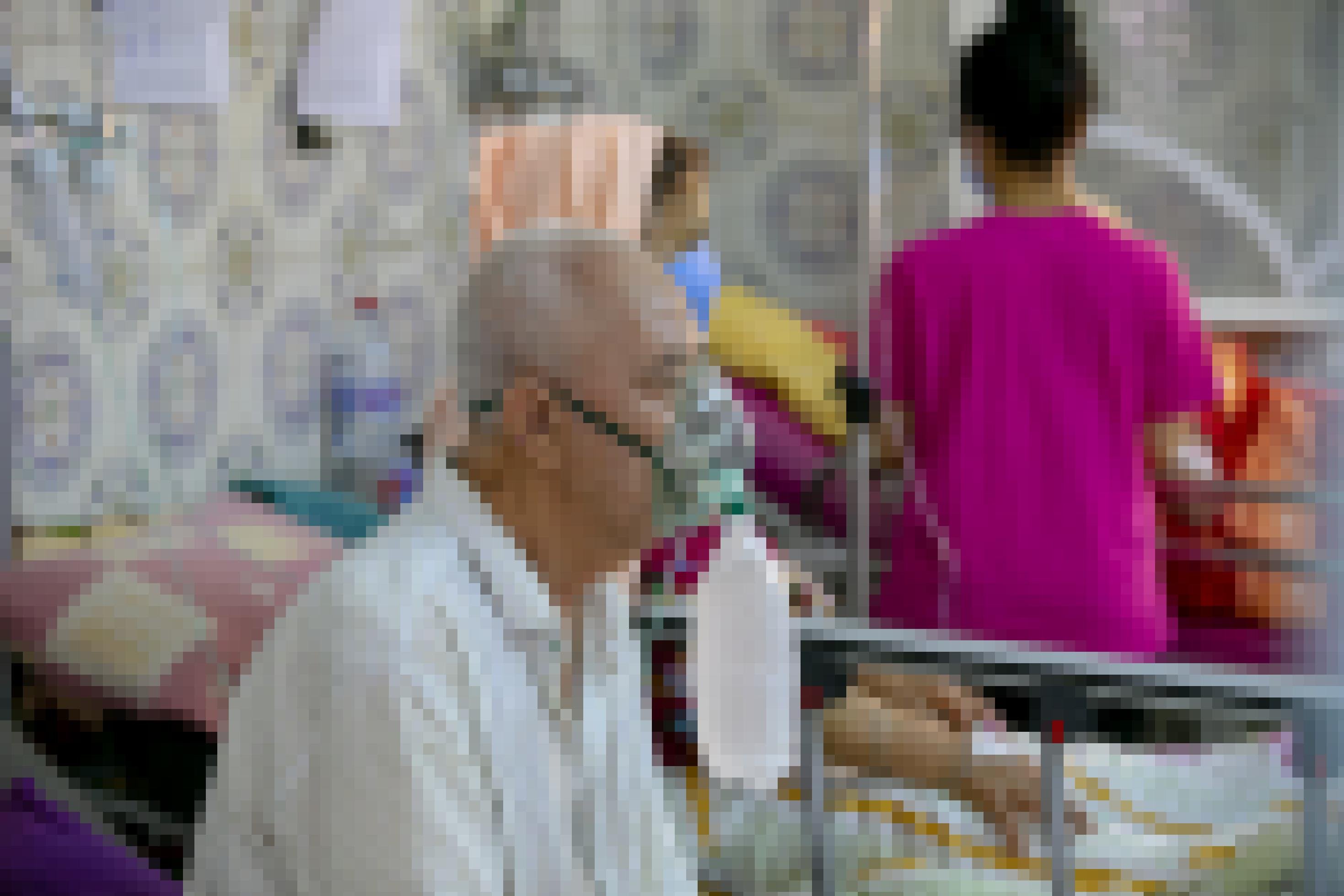 Ein älterer Mann, der mit Sauerstoff versorgt wird, sitzt in einem Krankenhausbett. Im Hintergrund sind weitere Personen zu erahnen.