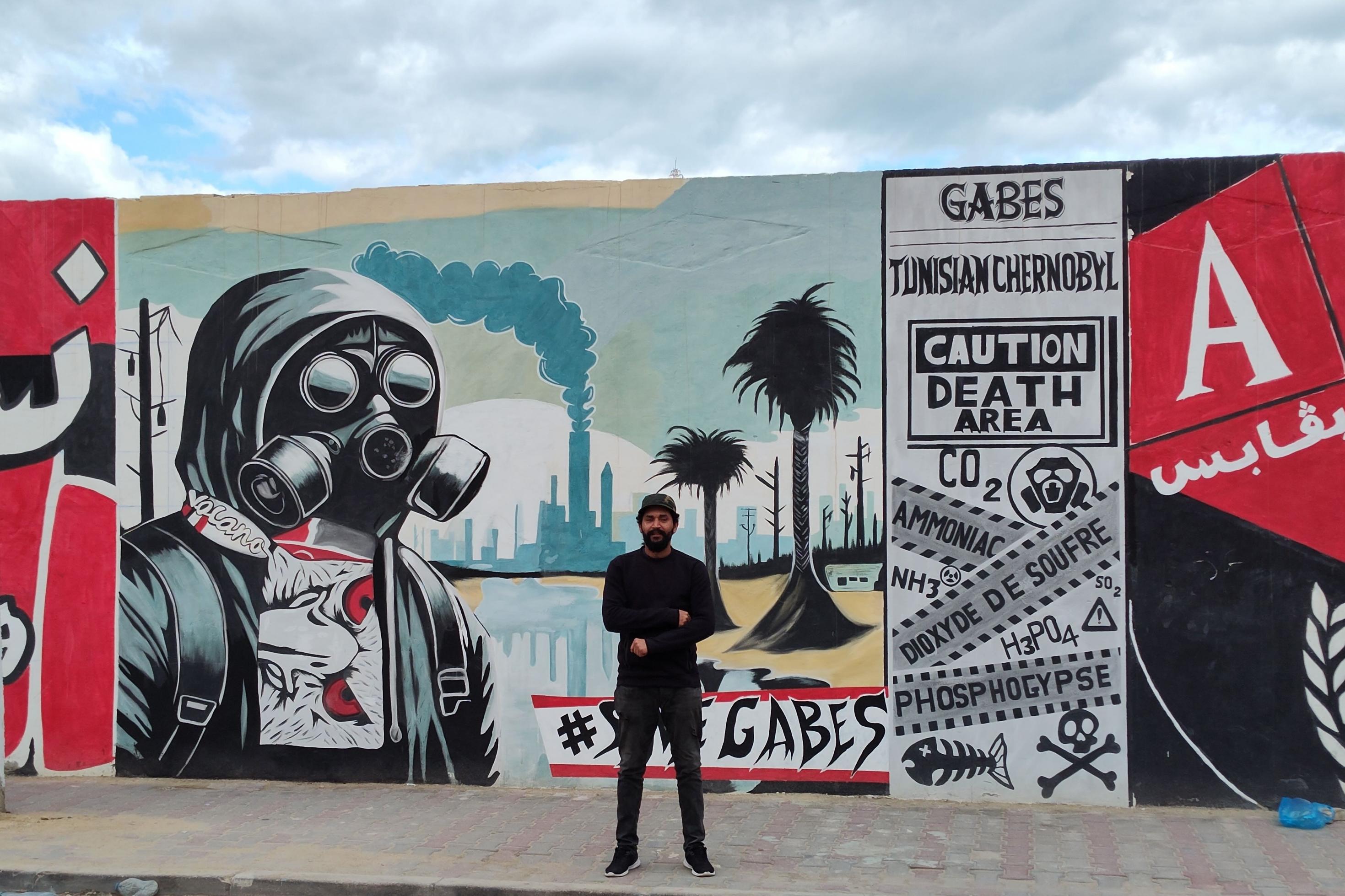 Ein junger Mann in schwarzer Kleidung steht mit verschränkten Armen vor einem Grafitti, auf dem eine Person. mit Gasmaske und Industrieanlagen zu sehen sind.