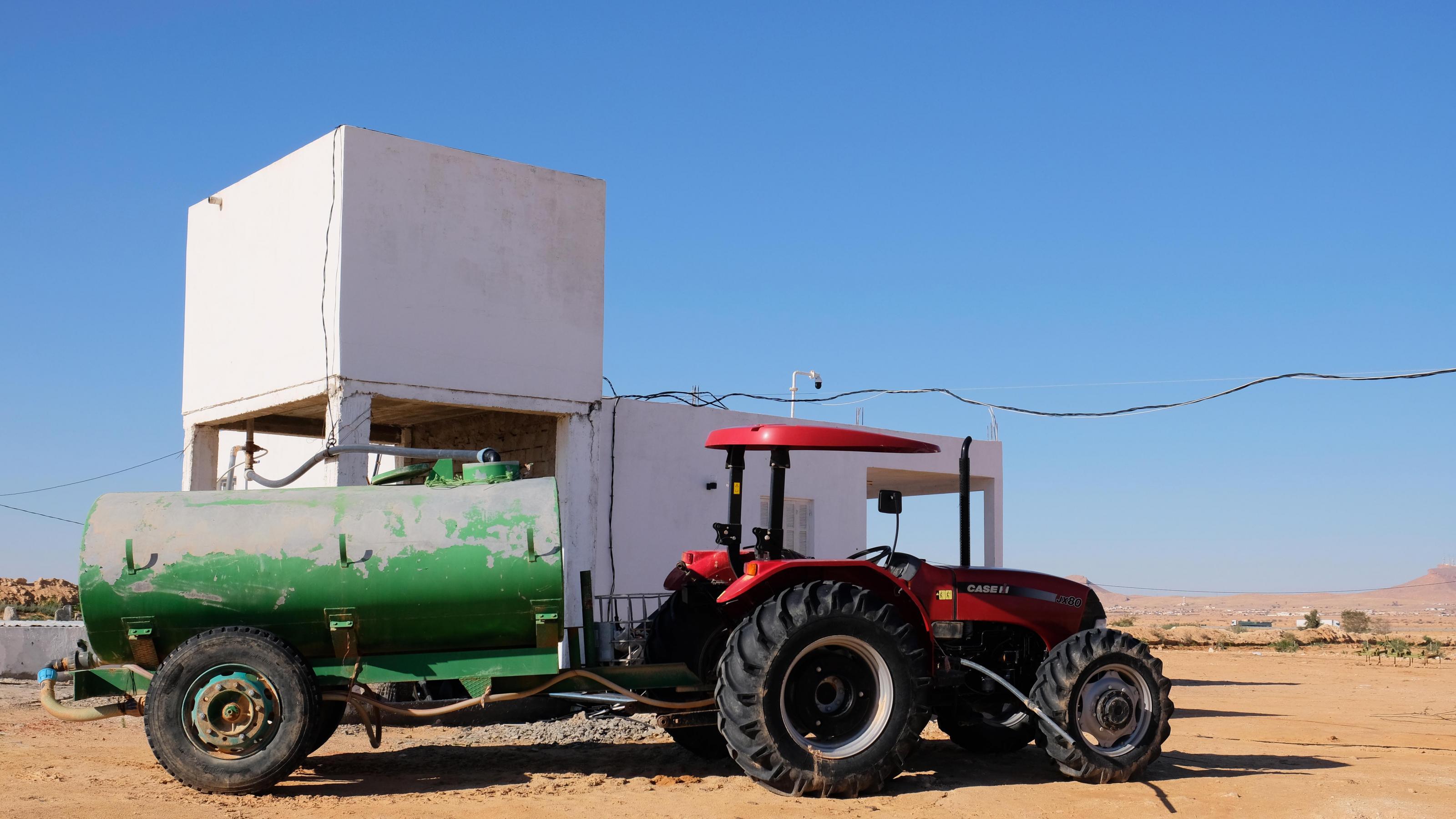 Ein weißer, quadratischer Wasserturm, vor dem ein roter Traktor mit grünem Anhänger steht.