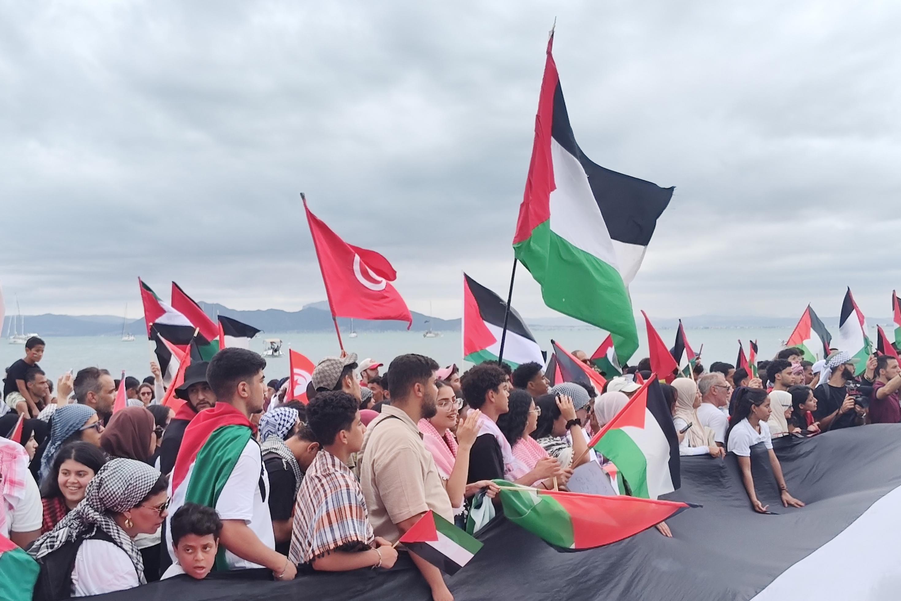 Eine Menschenmenge steht am Strand und schwenkt palästinensische und tunesische Fahnen.