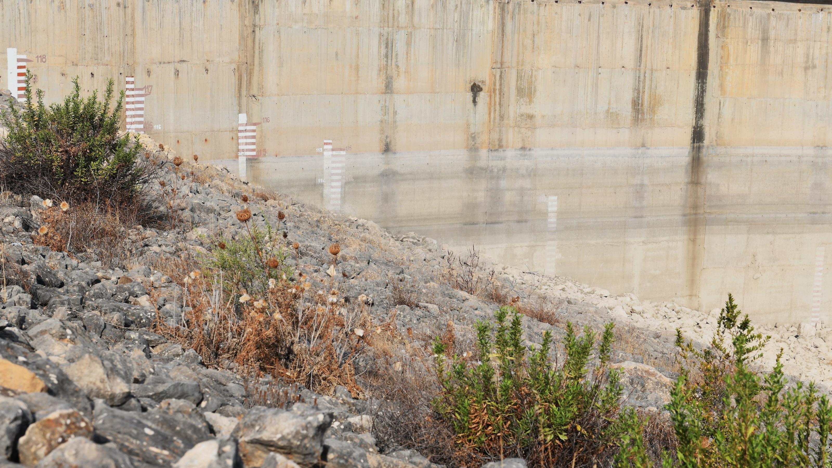 An der Mauer eines Staudamms sind die Spuren des Wasserstands vergangener Jahre und verblichene Höhenangaben zu sehen. Im Vordergrund wachsen Büsche auf Kies.
