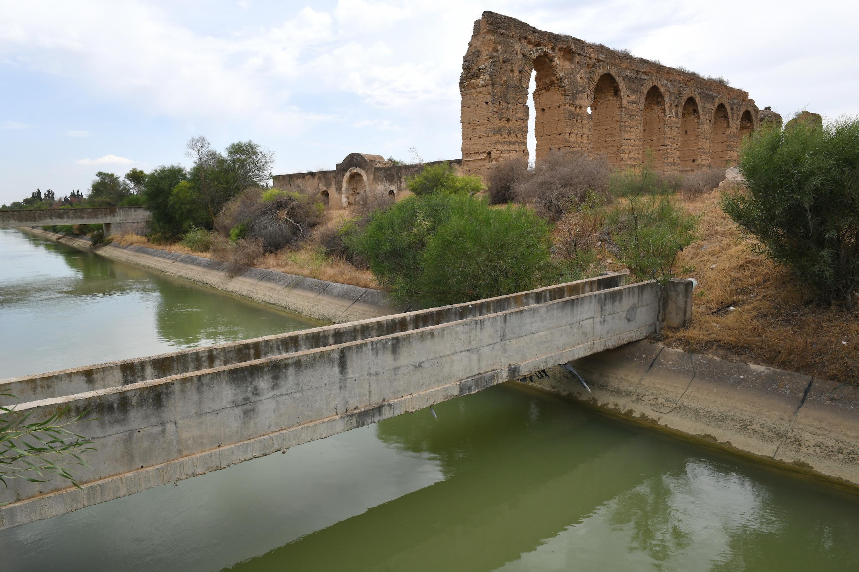 Im Hintergrund sind Ruinen eines römischen Aquädukts zu sehen, im Vordergrund der verschmutze Kanal Medjerda – Cap Bon