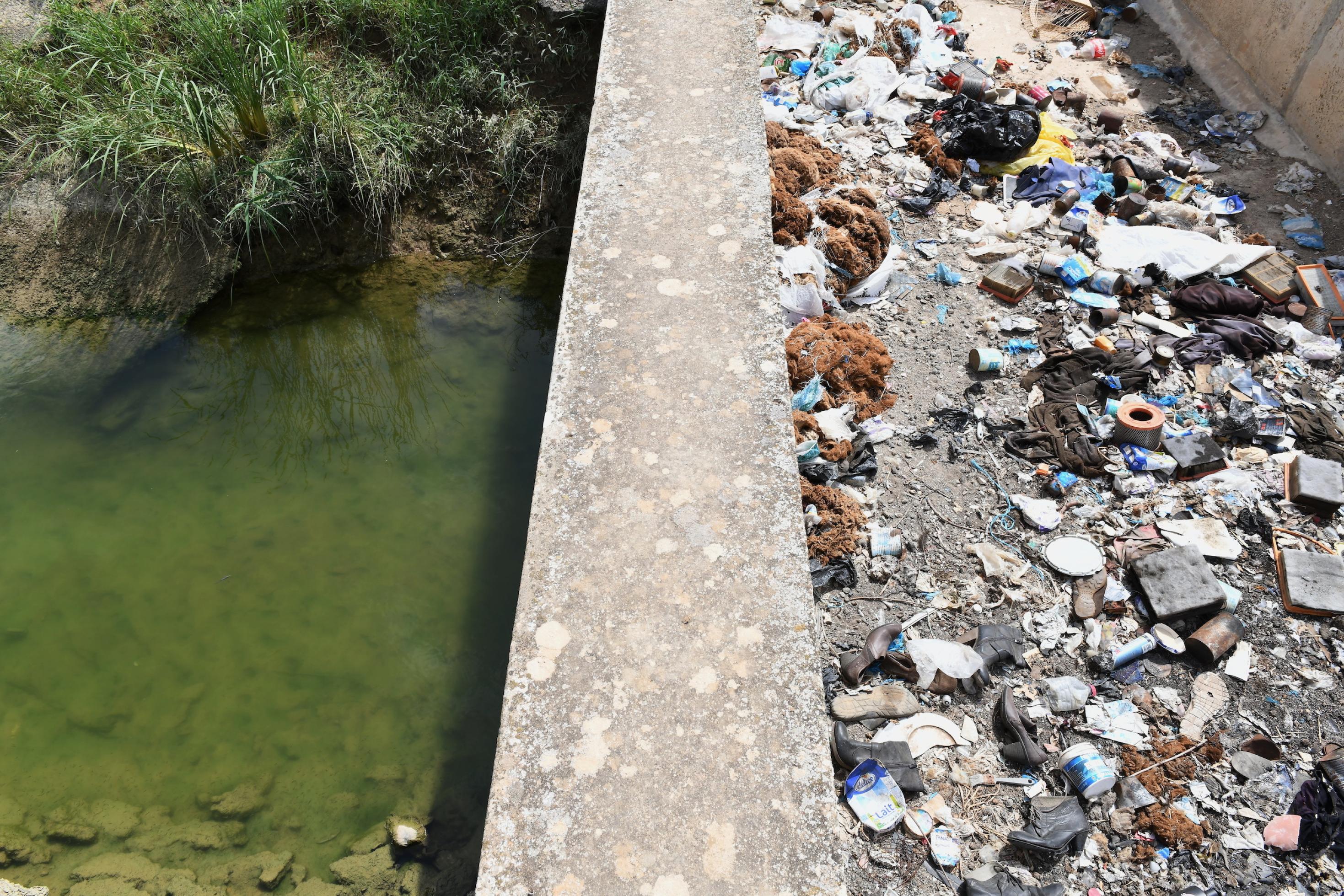 Trübes Wasser in einem Kanal, über den eine mit Müll überfüllte Brücke führt