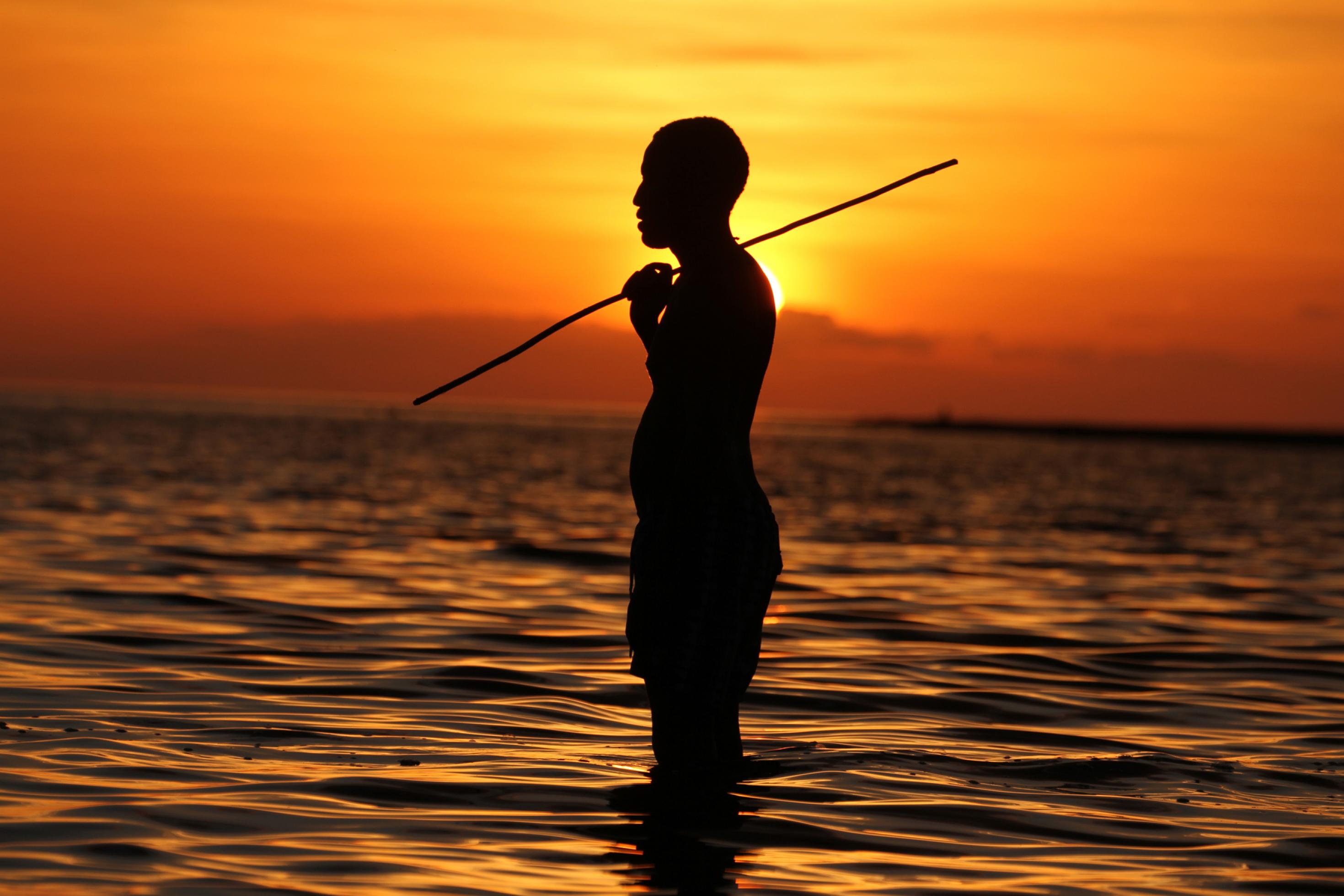 Ein afrikanischer Mann steht vor der untergehenden Sonne knietief im Wasser eines Sees, das leichte Wellen wirft. Er ist von der Seite zu sehen, hat einen Stock über der Schulter und schaut nach links. Durch das Gegenlicht ist nur die Silhouette des Mannes zu erkennen. Der Horizont leuchtet in goldgelben bis rötlichen Farben.