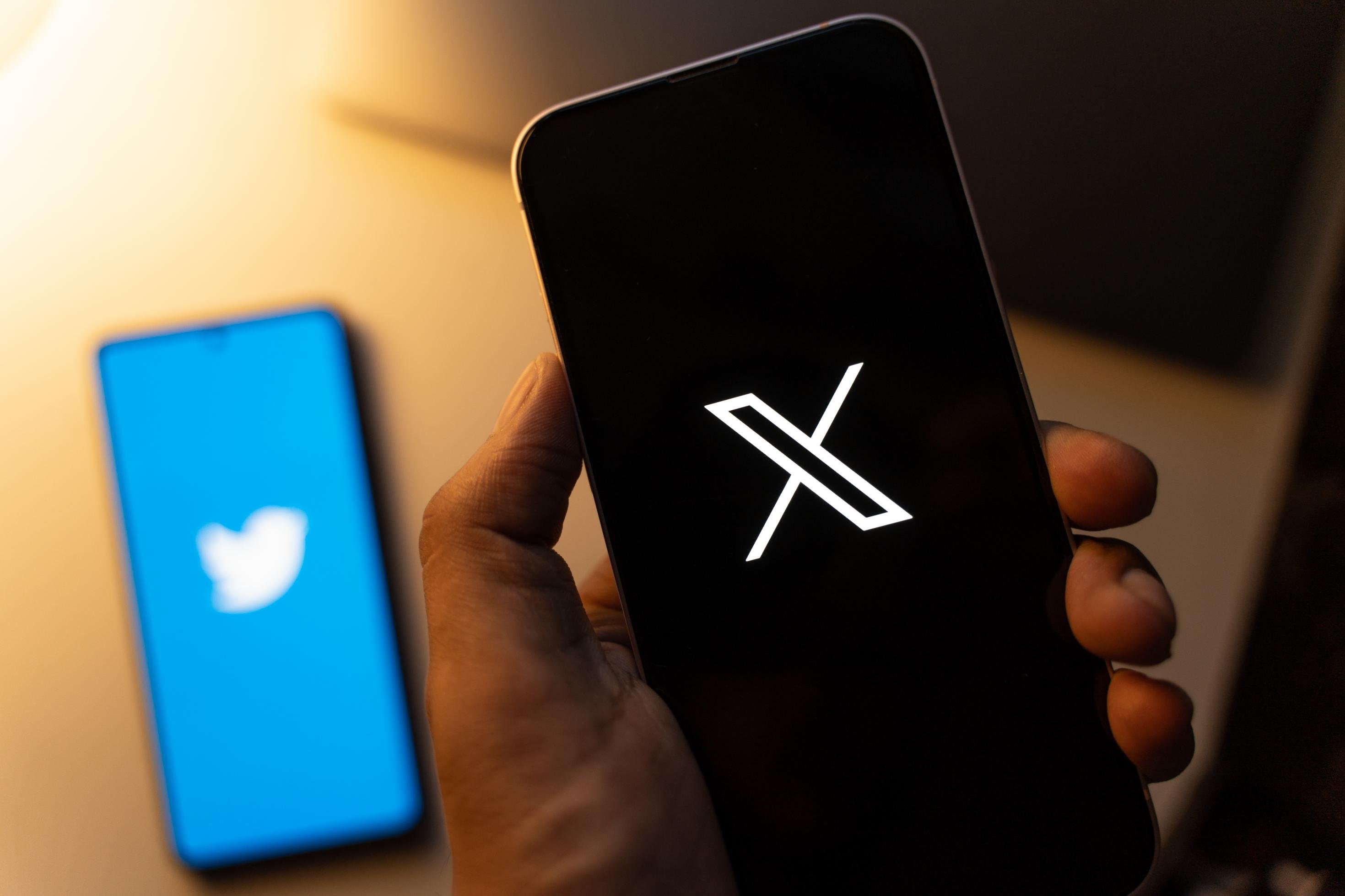 Man sieht eine Hand, die ein Smartphone in der Hand hält mit dem Symbol X. Ein zweites Smartphone liegt auf einem Tisch, dort ist der bekannte Twittervogel auf blauem Hintergrund zu sehen.