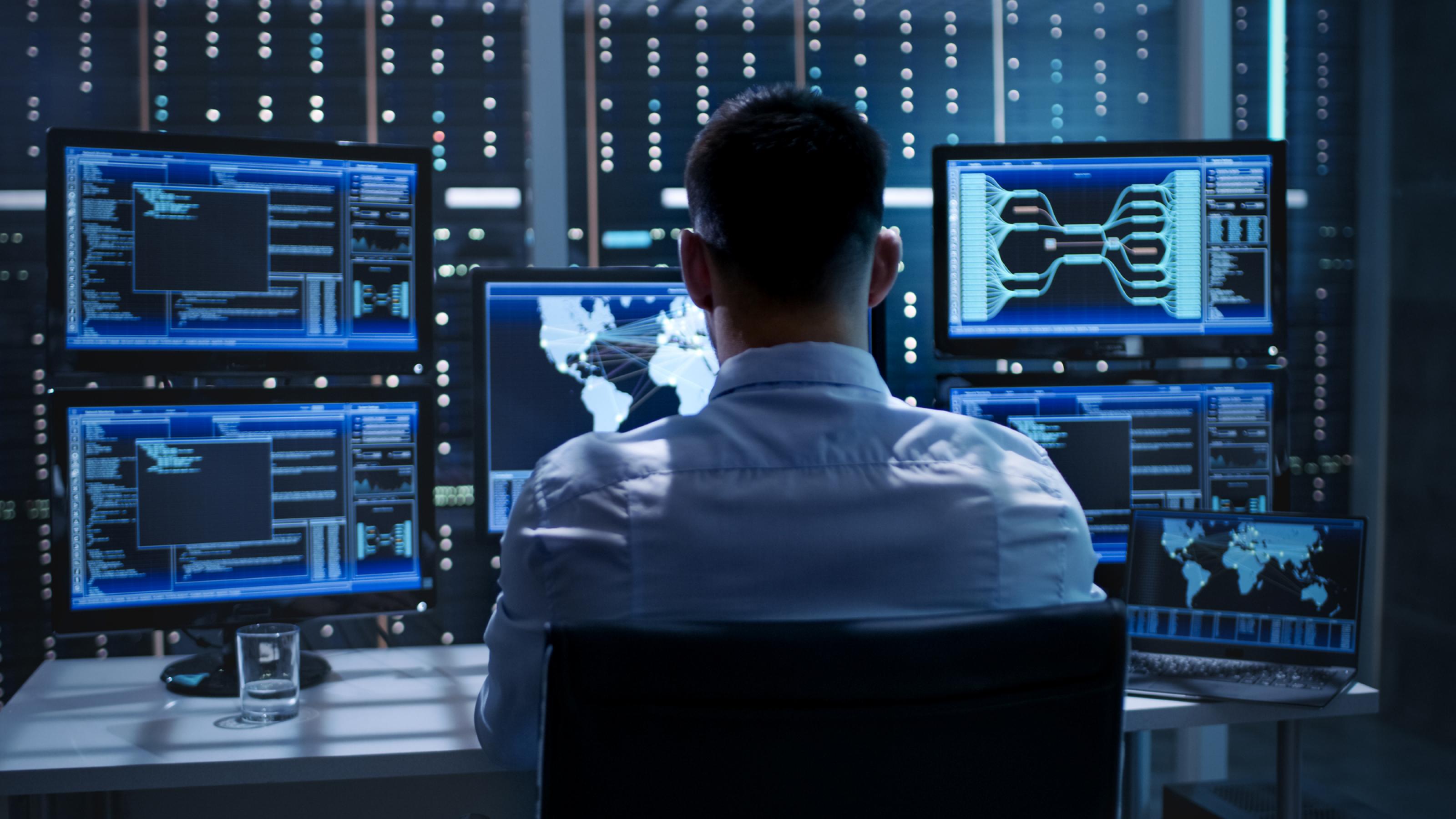 Ein Mann sitzt in einem Büro vor vielen Bildschirmen, die blau leuchten