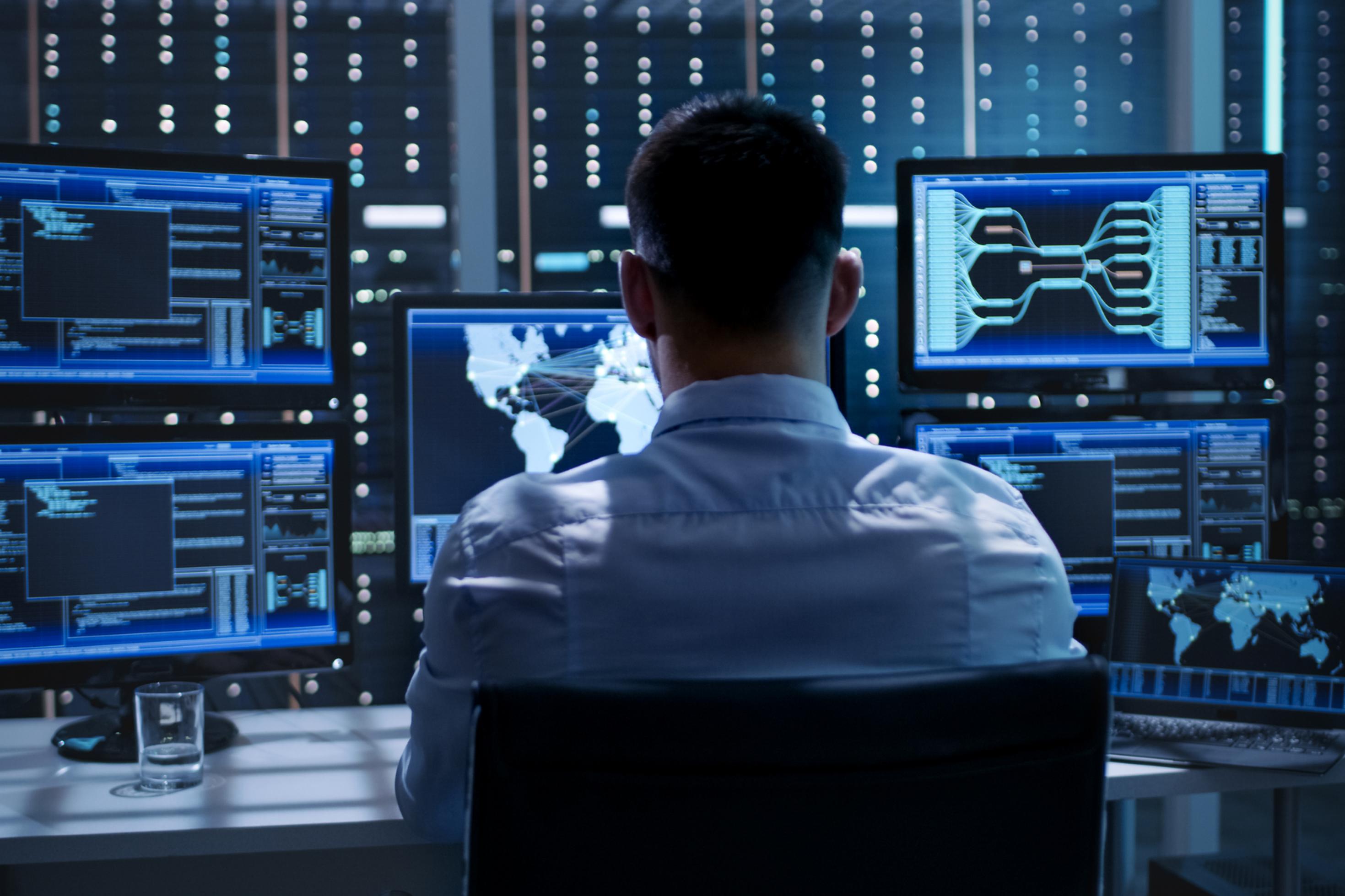 Ein Mann sitzt in einem Büro vor vielen Bildschirmen, die blau leuchten