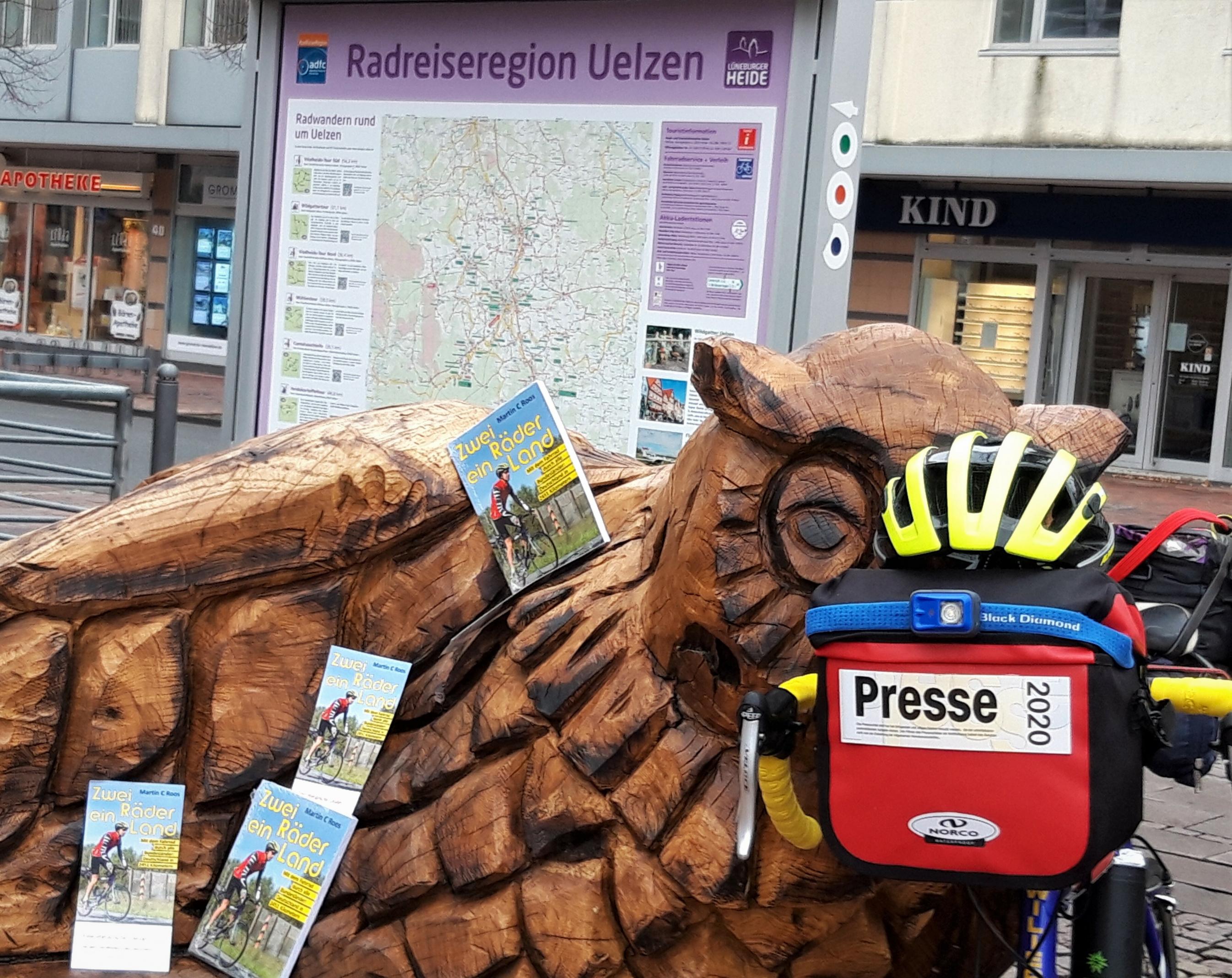 Vor einer Tafel mit der Aufschrift „Radreiseregion Uelzen“ sind die Flyer des Buchs „Zwei Räder, ein Land“ auf einer Holzbank verteilt.