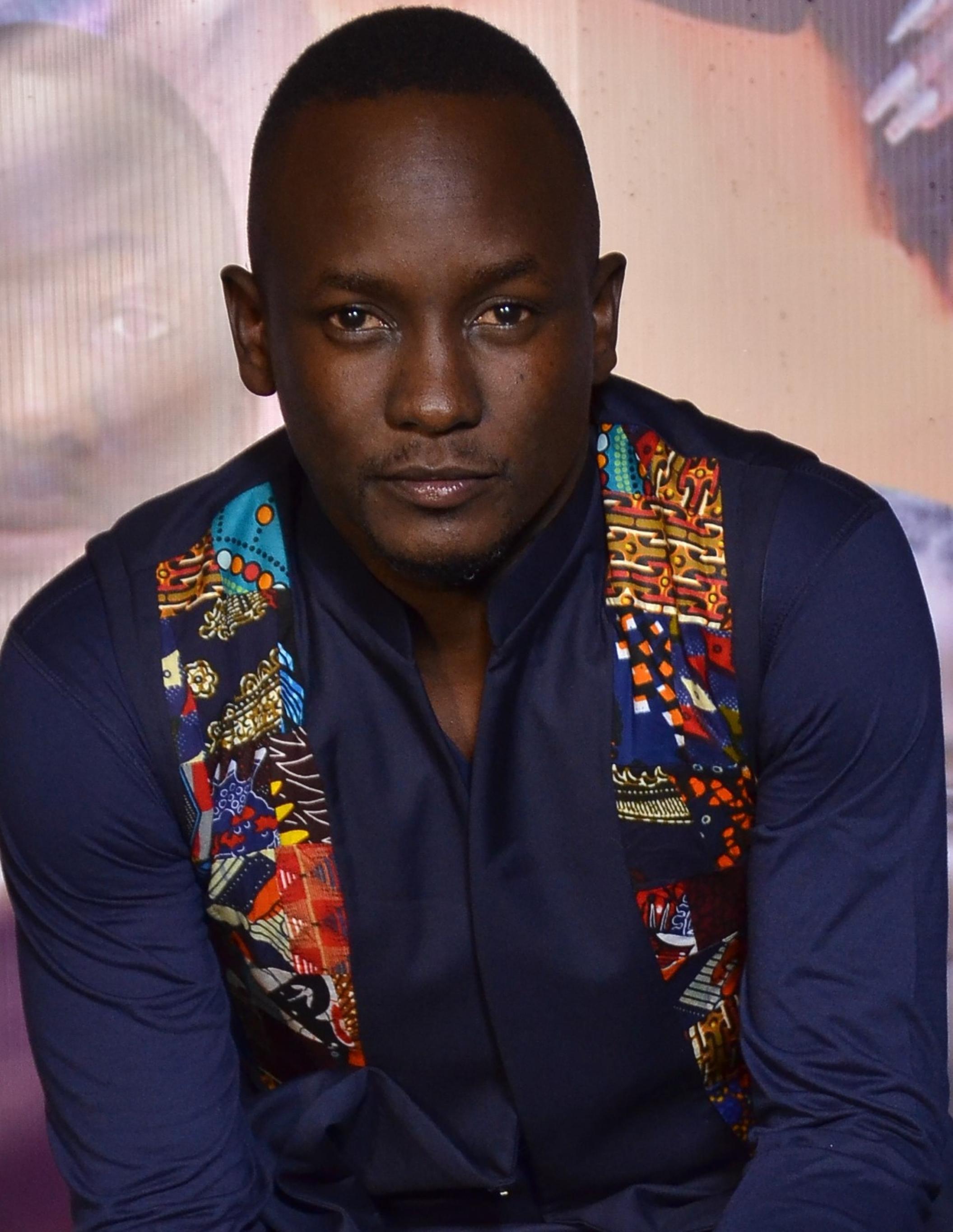 Abaas Mpindi schaut den Betrachtenden schräg von unten an. Er trägt ein dunkelblaues Hemd und eine Weste mit buntem, afrikanischem Muster.