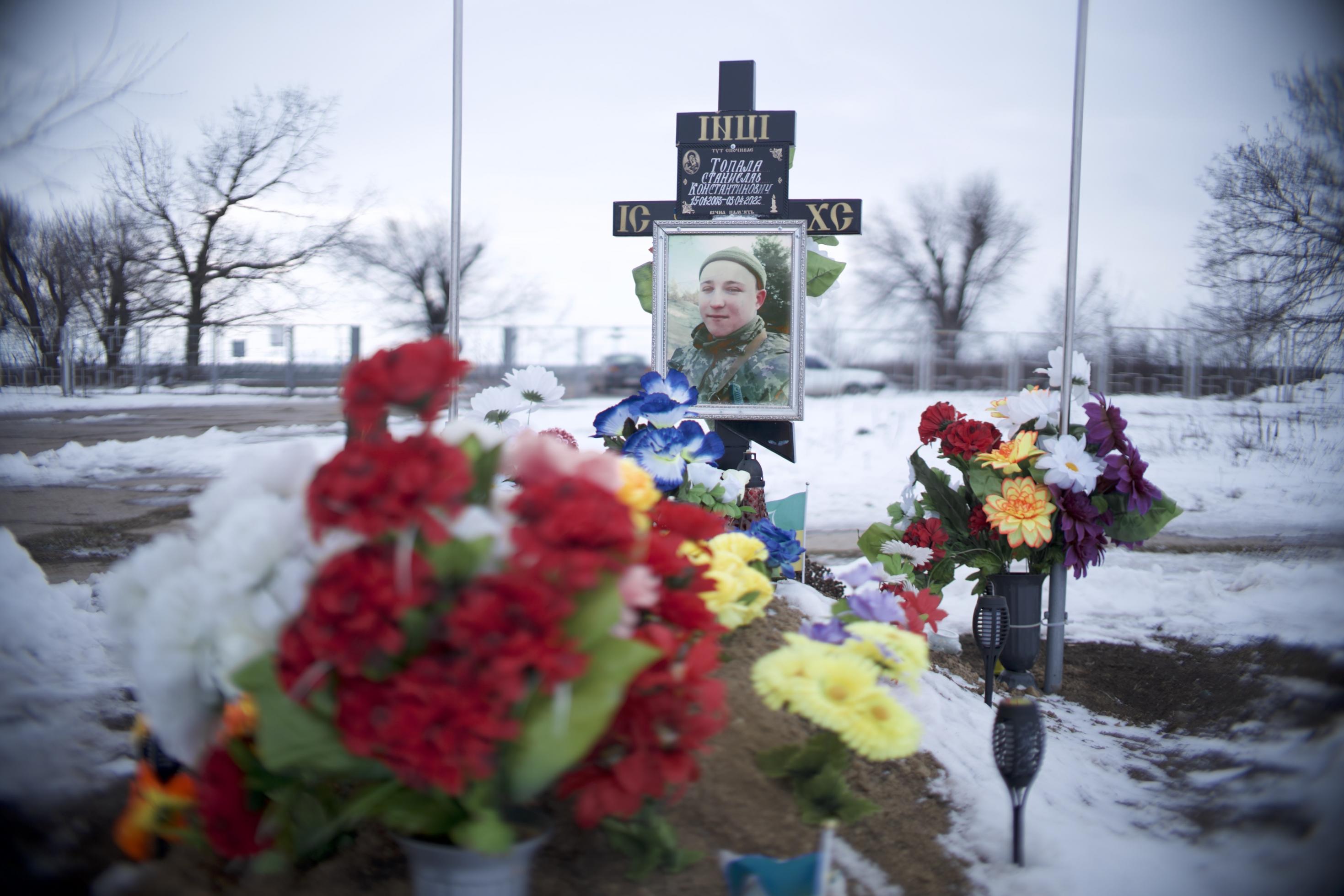 Hinter Blumen erscheint ein Kreuz mit einem Bild eines im Ukrainekrieg getöteten Soldaten