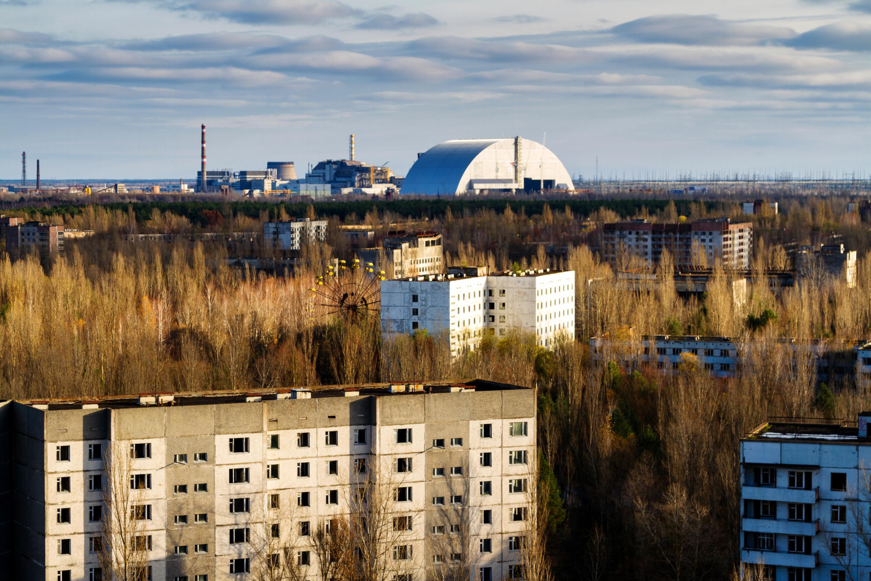 Blick von hoher Warte auf das Nukleargelände, im Vordergrund leerstehende Hochhäuser, dann ein Riesenrad in einem Wald, dann der Sarkophag mit dem explodierten Reaktor, dahinter Wolken