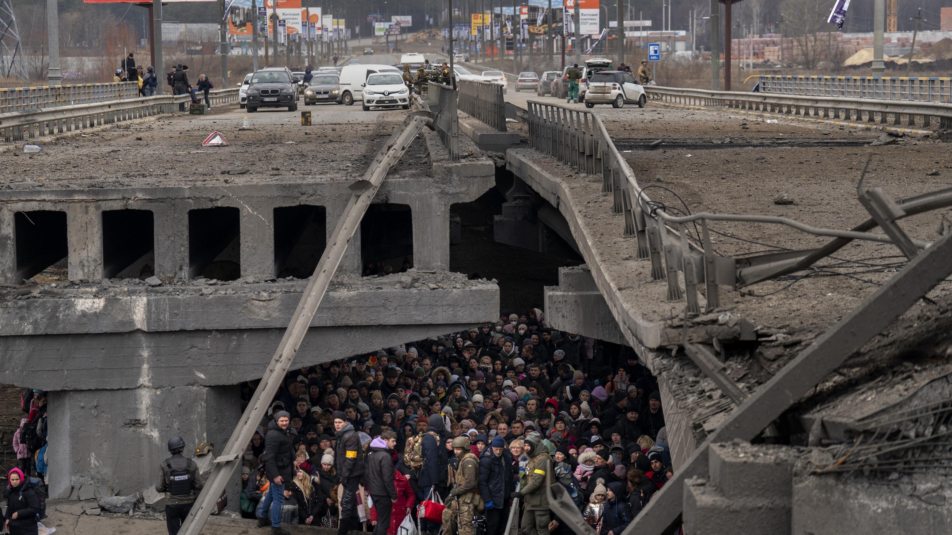 Im Dunkel unter der zerstörten Brücke wartet eine Menschenmenge, bewacht von zwei ukrainischen Soldaten.