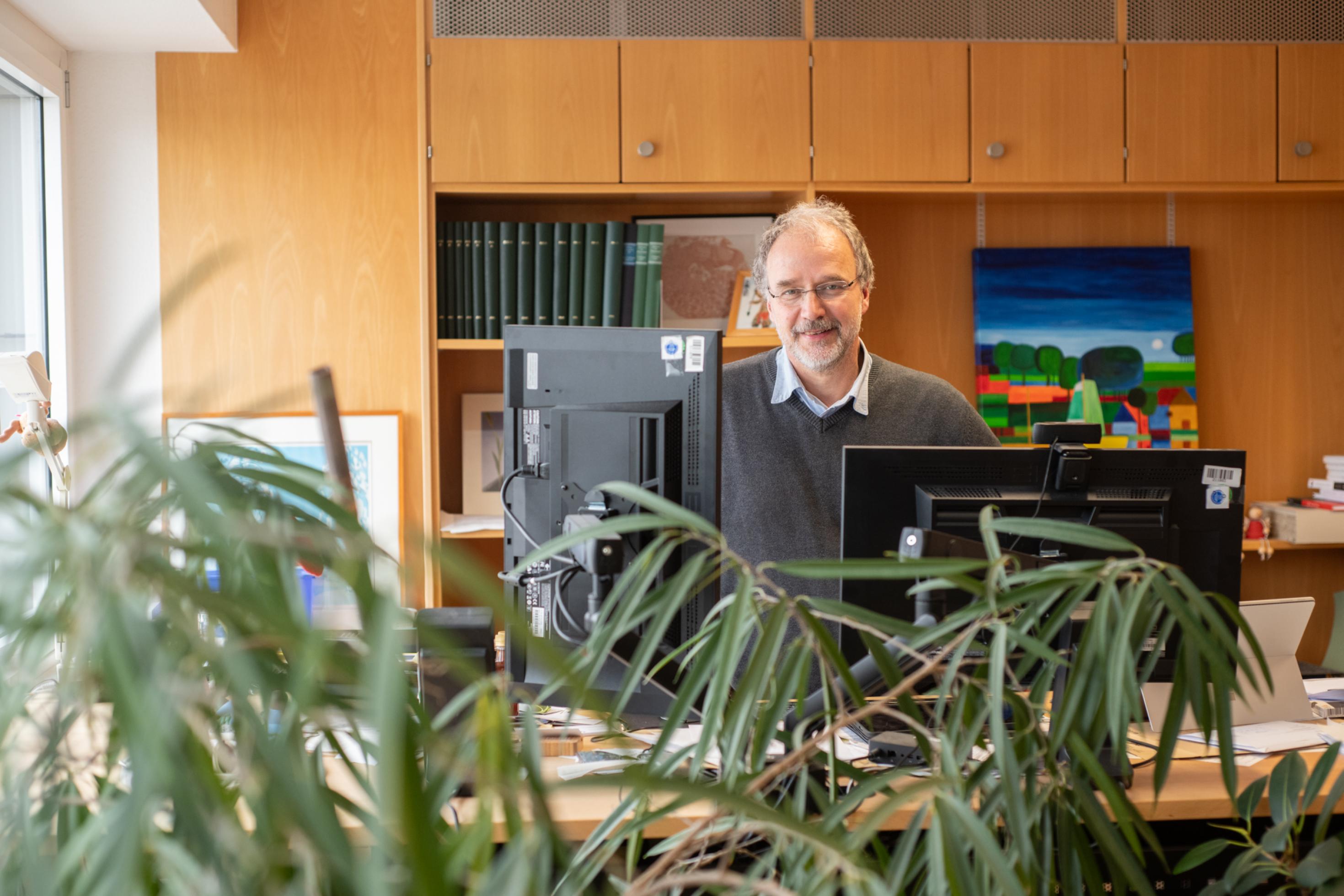 Ein Mann mit grauem Haar und Brille steht hinter einem Schreibtisch mit zwei Monitoren, davor mehrere große grüne Pflanzen.