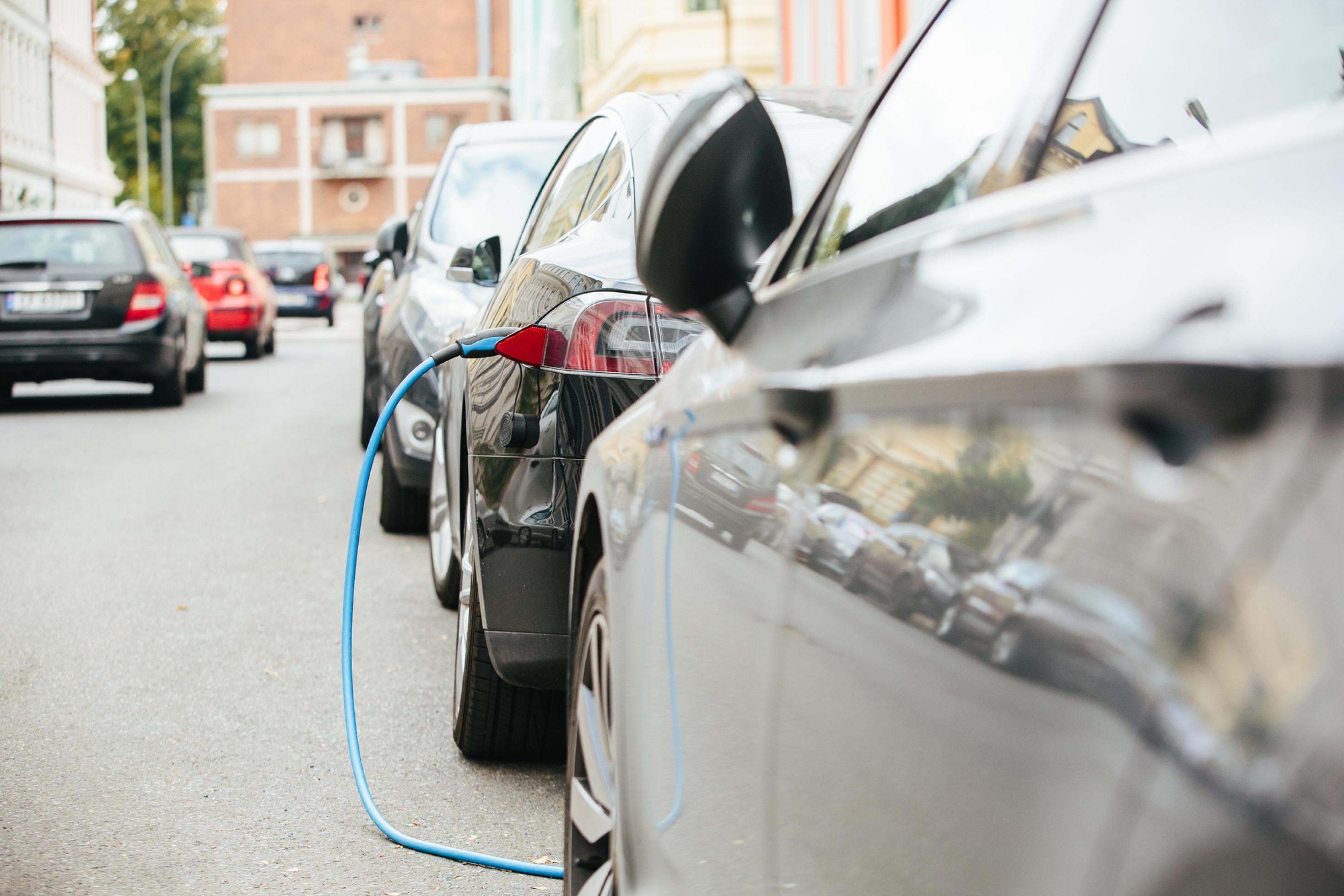 Das Bild zeigt eine Straße voll mit fahrenden und parkenden Autos, von denen eines ein Elektroauto ist und am Straßenrand mit einem Stromkabel neu aufgeladen wird.