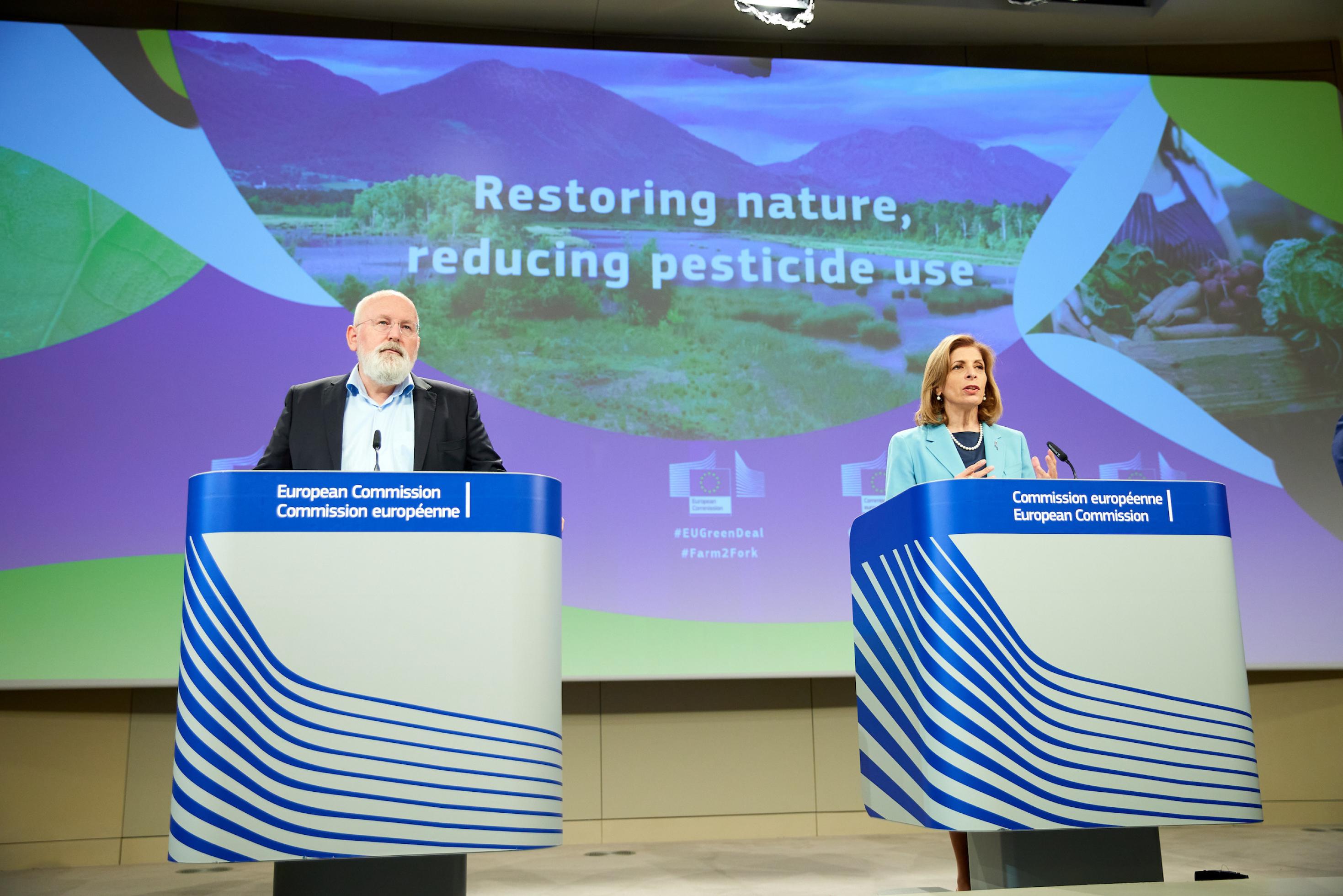 Der glatzköpfige Timmermans und EU-Kommissarin Stella Kyriakides stehen an Pulten vor einem großen Bildschirm mit der Aufschrift „Restoring Nature, reducing pesticides“